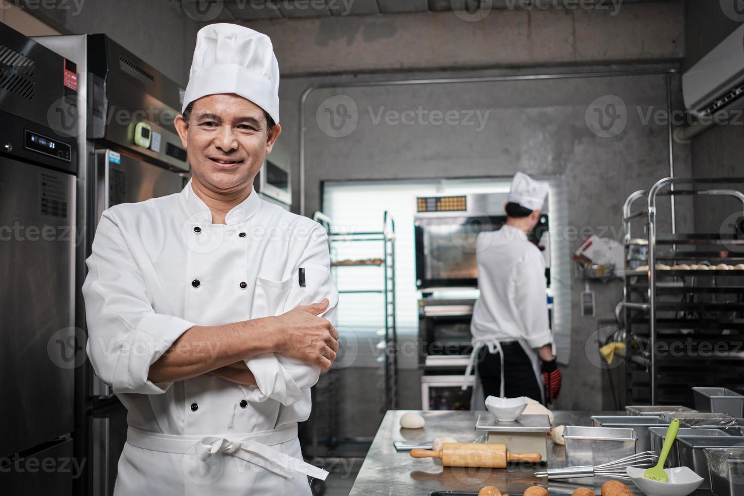 Professional senior Asian male chef in white cooking uniform, food occupation looks at camera, arms crossed, cheerful smile, expertise in commercial pastry culinary jobs, catering restaurant kitchen. photo