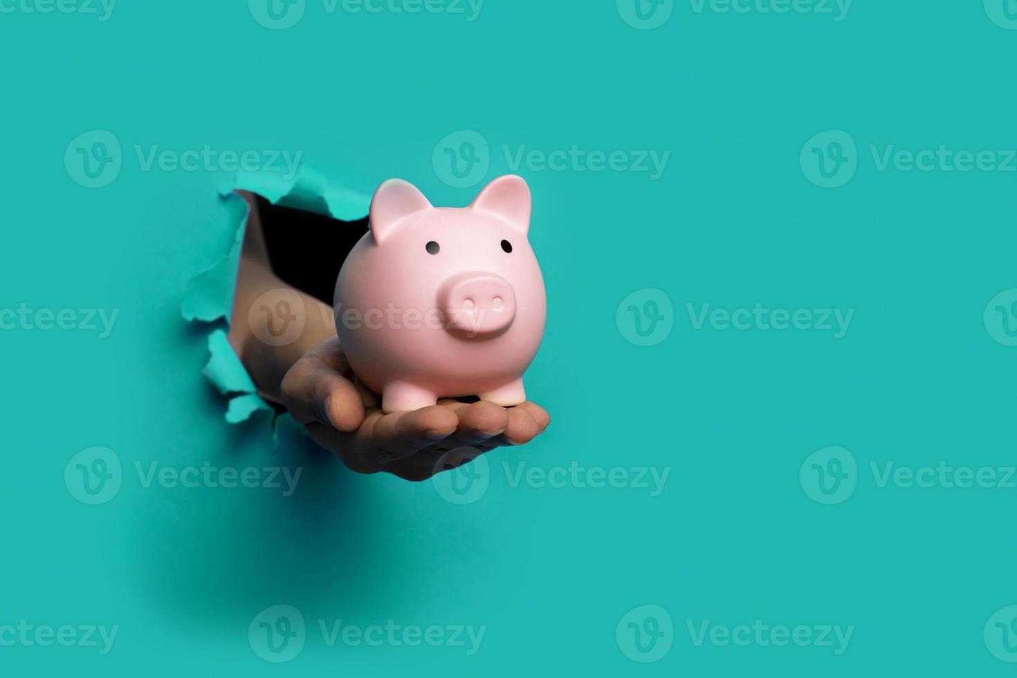 hand holds a piggy bank blue background out of a torn hole in a paper background photo