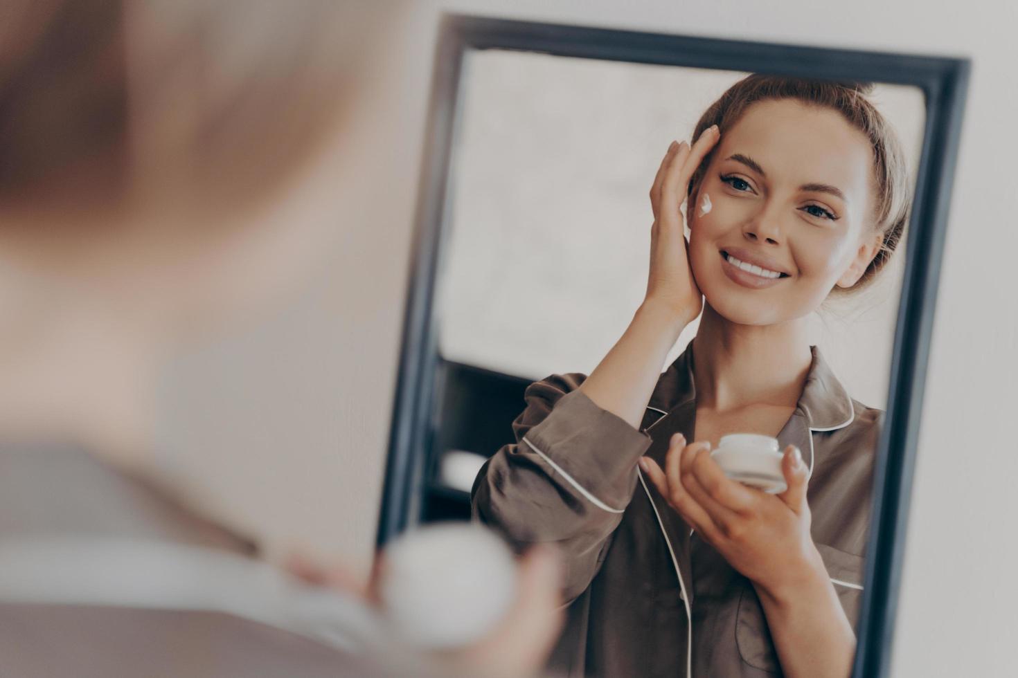 mujer joven positiva y feliz sonriendo mientras aplica crema facial foto