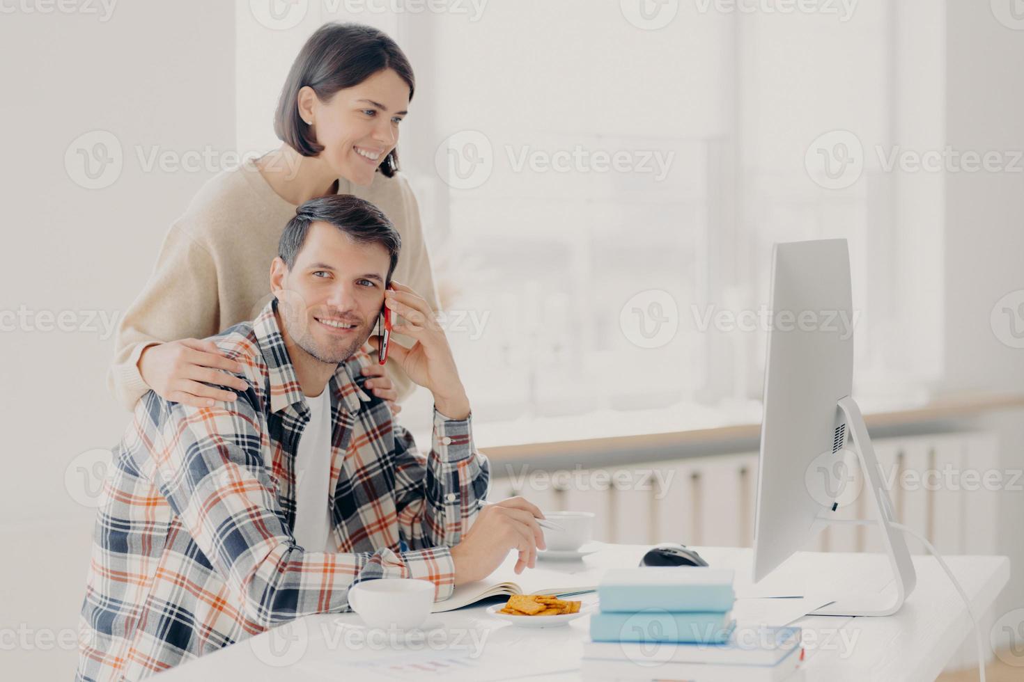 Happy husband and wife do paperwork together, pay taxes online look at screen computer, pose at desktop with books and papers, drink coffee and have lunch, make telephone call via smartphone photo