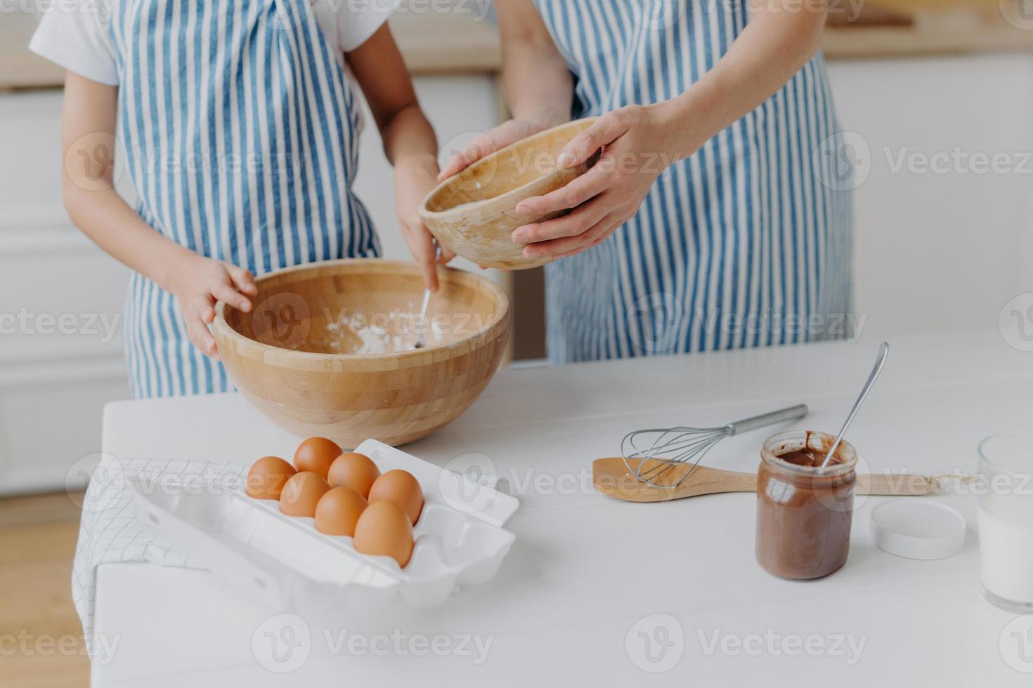 Cropped image of mothers and daughters hands mixing ingredients to prepare dough and bake tasty pastry, stand near kitchen table with eggs, melted chocolate in glass, wear striped blue aprons photo