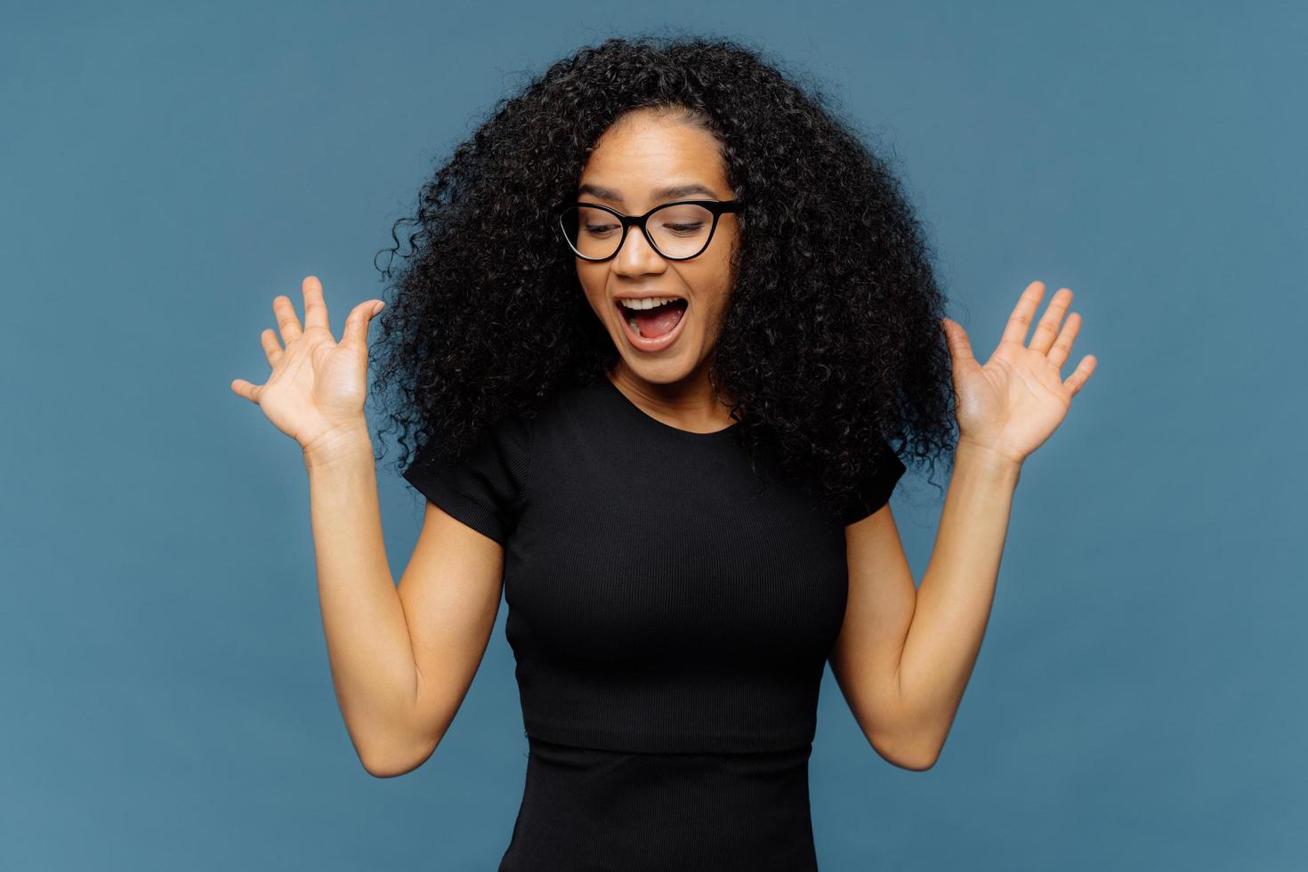 Slim overemotive dark skinned woman raises hands, opens mouth, gestures actively from positive emotions, focused down, dressed in casual black t shirt, isolated over blue background. Ethnicity concept photo