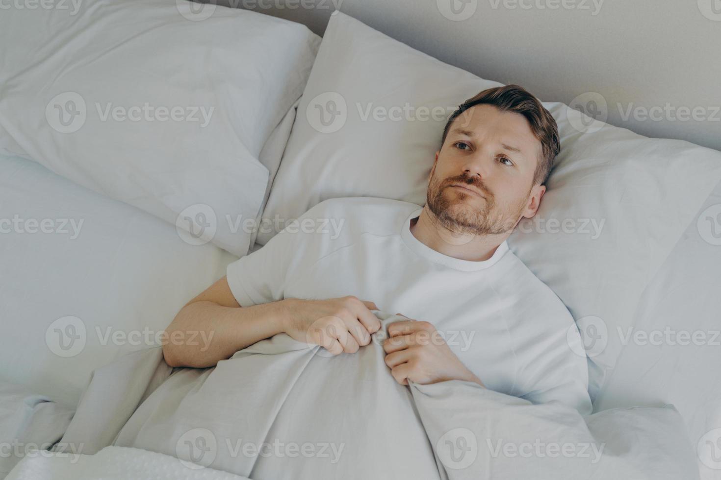 Lonely upset young bearded man lying in bed with open eyes photo
