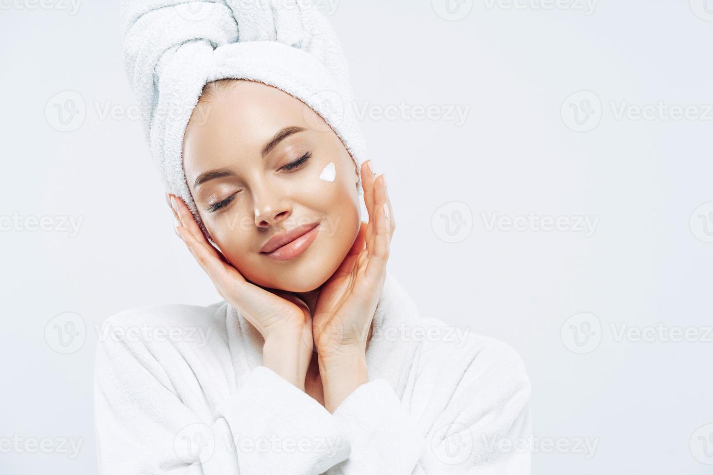 foto de estudio de una mujer relajada y hermosa con los ojos cerrados inclina la cabeza, toca la piel suavemente, aplica crema facial, usa una toalla de baño en la cabeza después de los procedimientos de spa, disfruta de tratamientos faciales, cuidado corporal