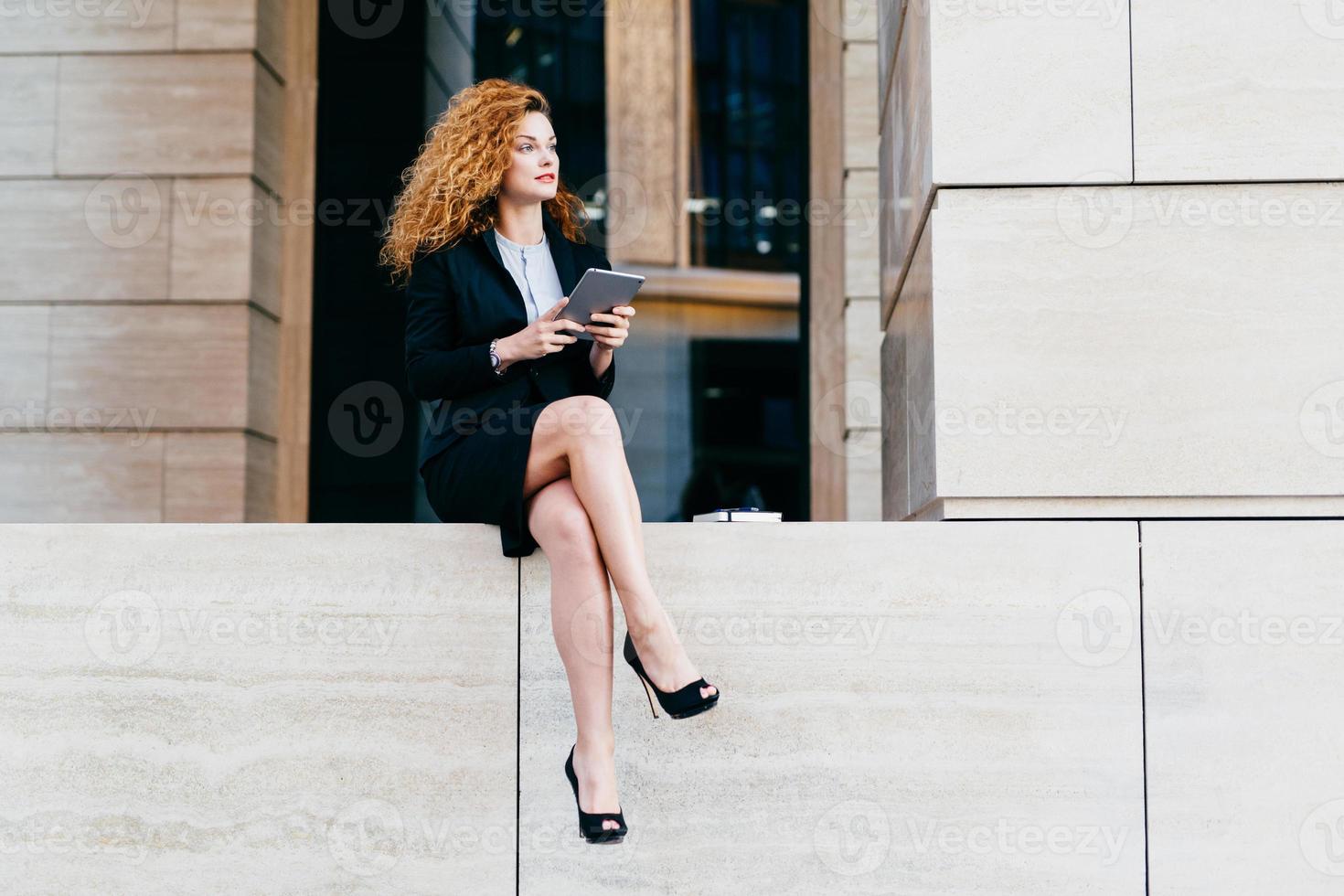 mujer de negocios delgada con cabello lujoso y ondulado, piernas delgadas, traje y zapatos negros elegantes, sosteniendo una tableta, mirando a un lado mientras descansa por un minuto después del trabajo duro en la oficina foto