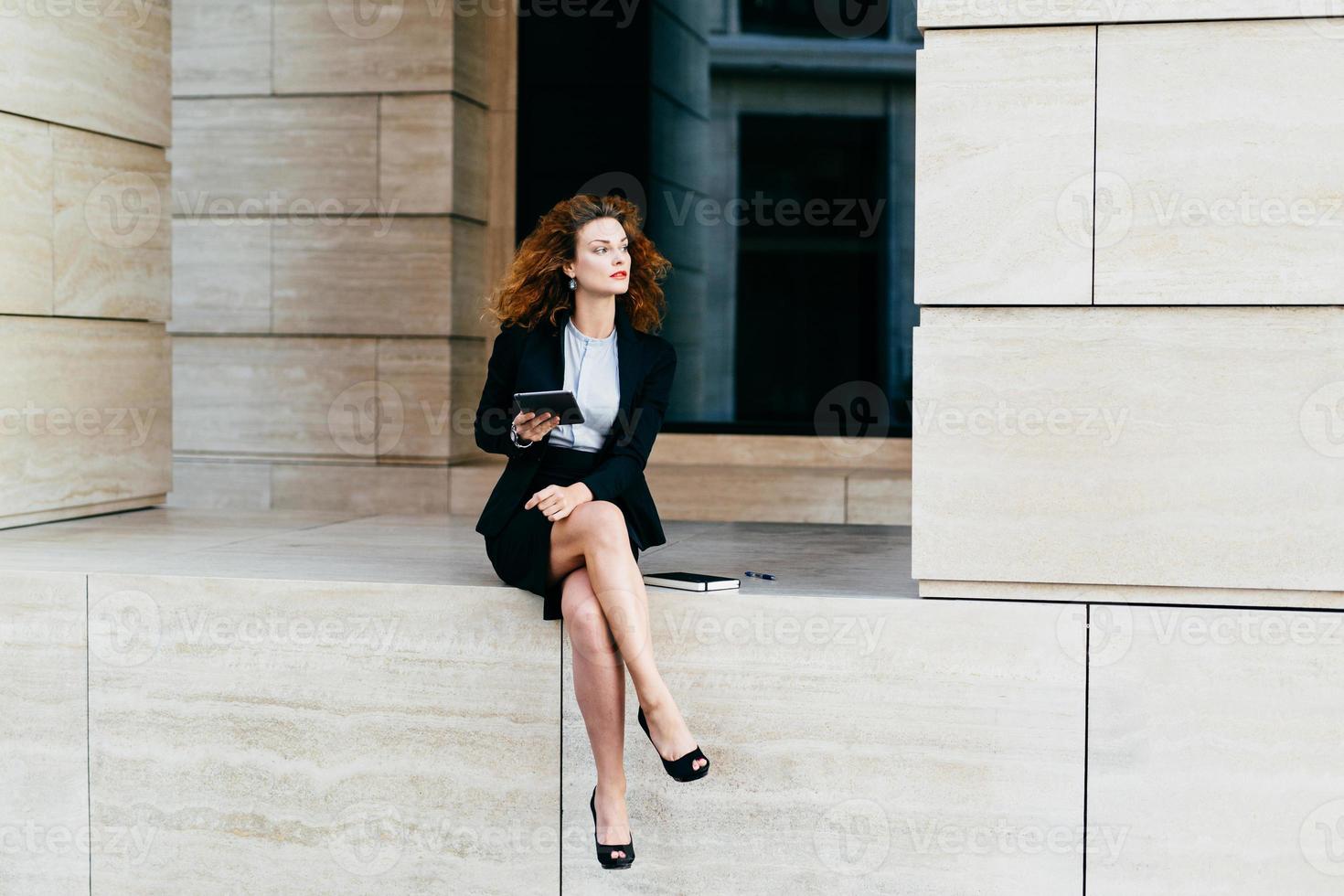 Female entrepreneur with slender legs, wearing black formal costume, sitting crossed legs, holding modern digital tablet computer, looking with pensive expression, planning her future partnership photo