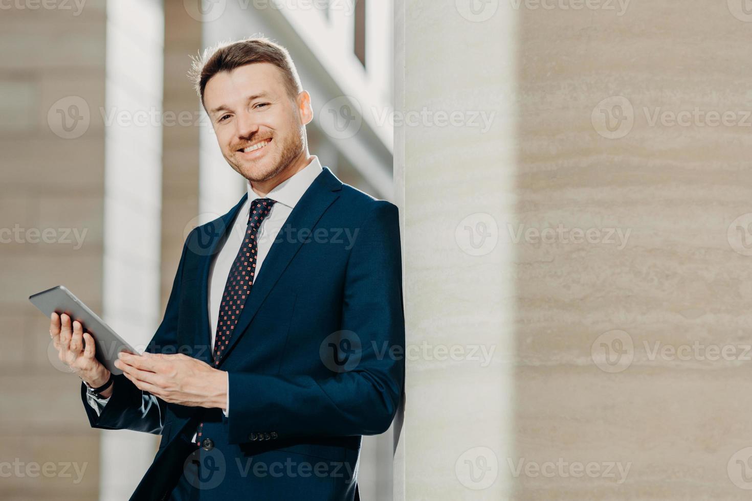 Portrait of stylish male entrepreneur has cheerful expression, holds digital tablet, reads financial news on web page, uses free internet connection, wears formal suit and tie. Career concept photo