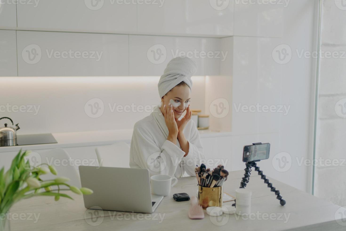 Female beauty blogger in bathrobe and towel on head recording video on smartphone for her vlog photo
