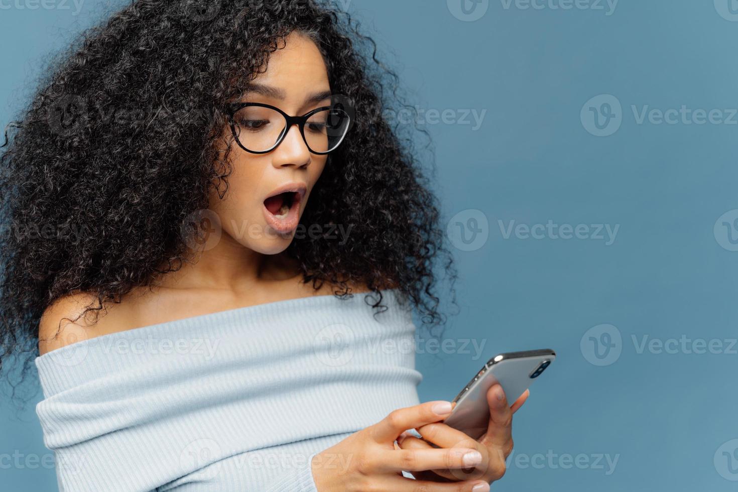 Shocked African American woman gots message from ex, gasps from surprise and excitement, reads stunning news on mobile phone, has curly bushy hair, wears jumper, isolated over blue studio wall photo