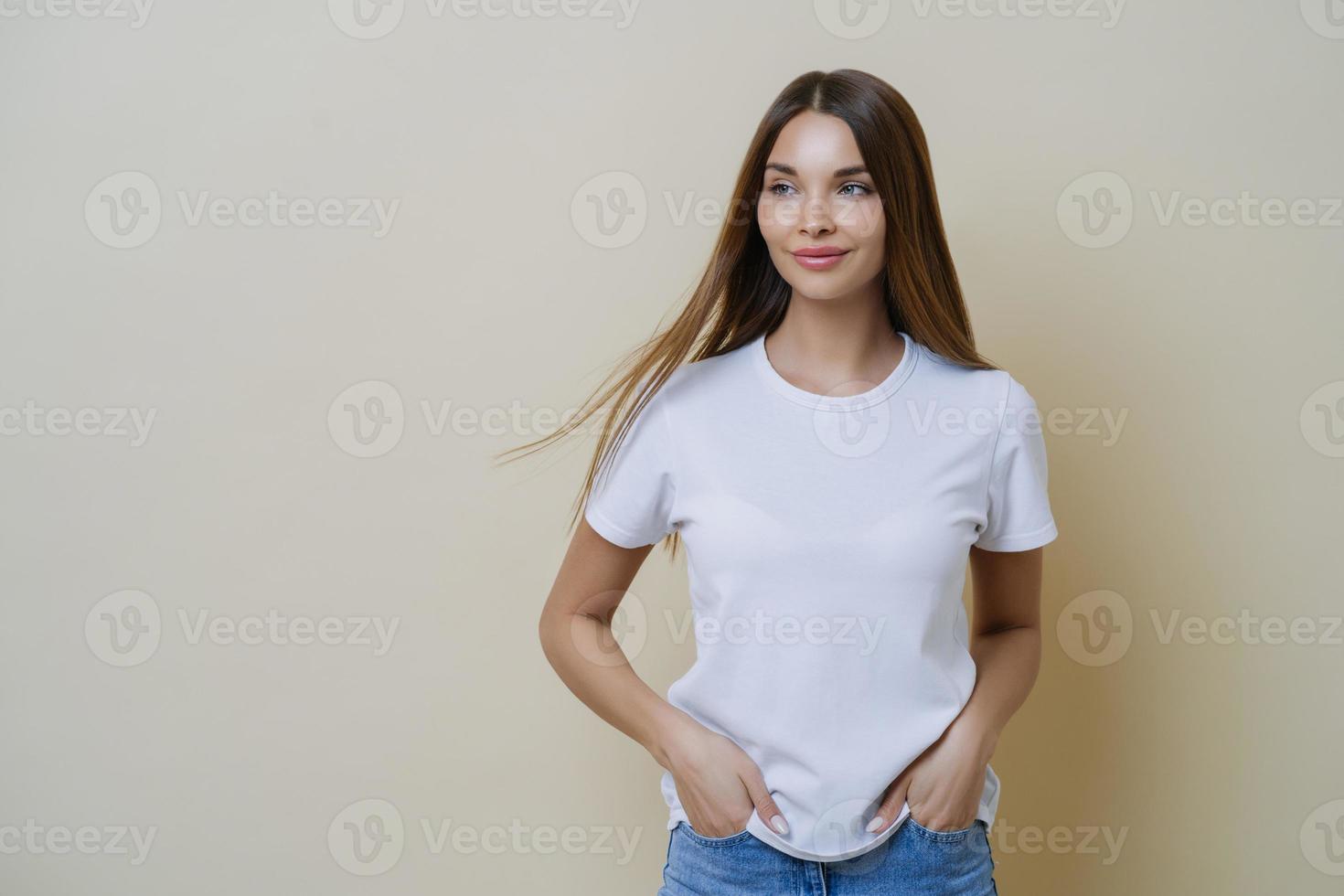 Thoughtful brunette woman looks aside, wears casual white t shirt, keeps hands in pockets, looks aside, isolated over beige background, has relaxed face expression, empty space for your promotion photo