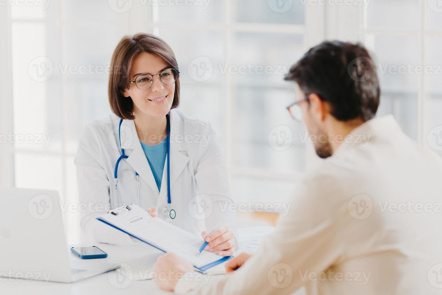 Young female doctor consults patient, shows information and prescription in clipboard, talk about medical checkup, pose at clinic, explains health insurance contract, read service agreement. photo