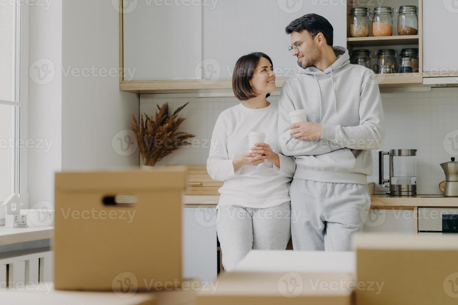 Image of cheerful woman and man have pleasant talk during coffee break, wear casual clothes, move in new apartment, pose with modern furnished kitchen, unpack cardboard boxes with household stuff photo