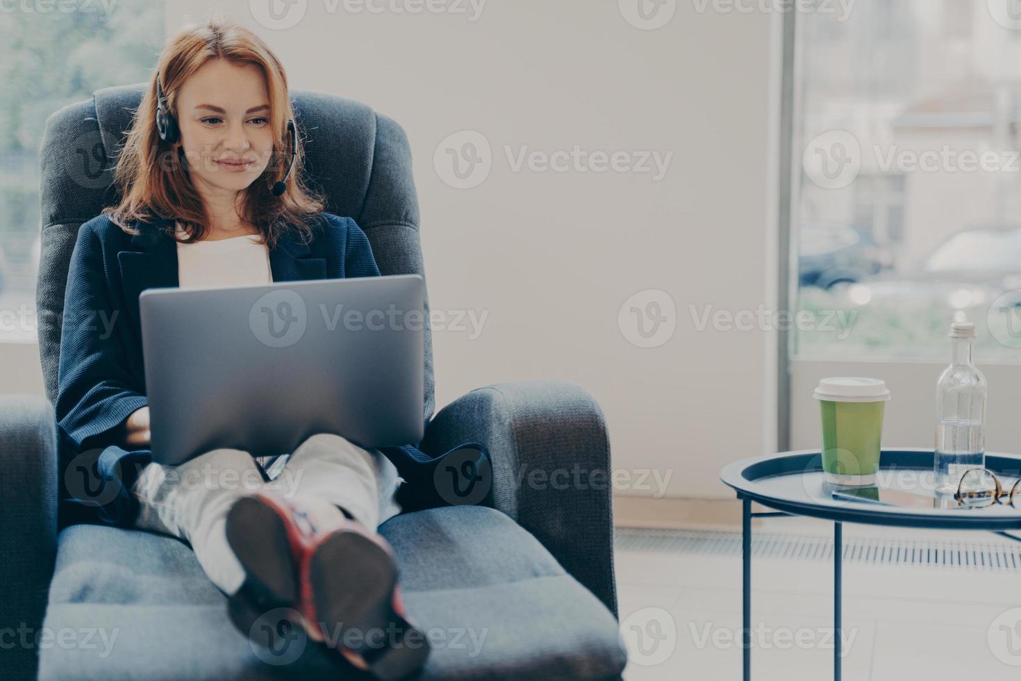 mujer pelirroja sonriente que trabaja con auriculares en una computadora portátil en una oficina moderna y abierta foto