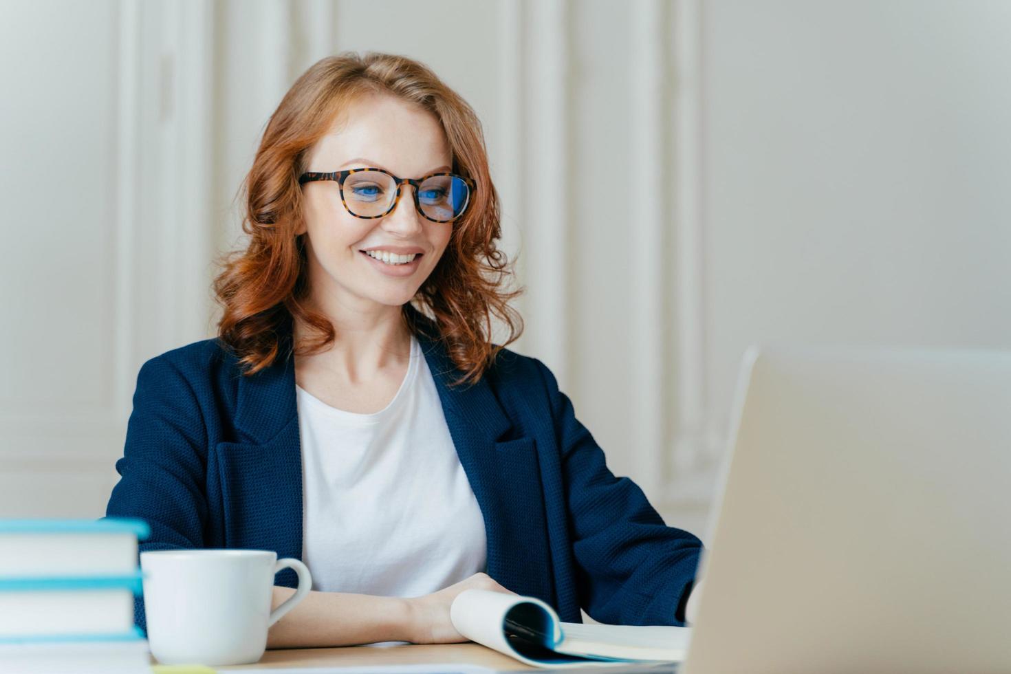 Glad lovely woman has ginger hair, positive smile, sits with laptop computer at desktop, happy to make video call or conference, communicates with investors from abroad, drinks aromatic coffee. photo