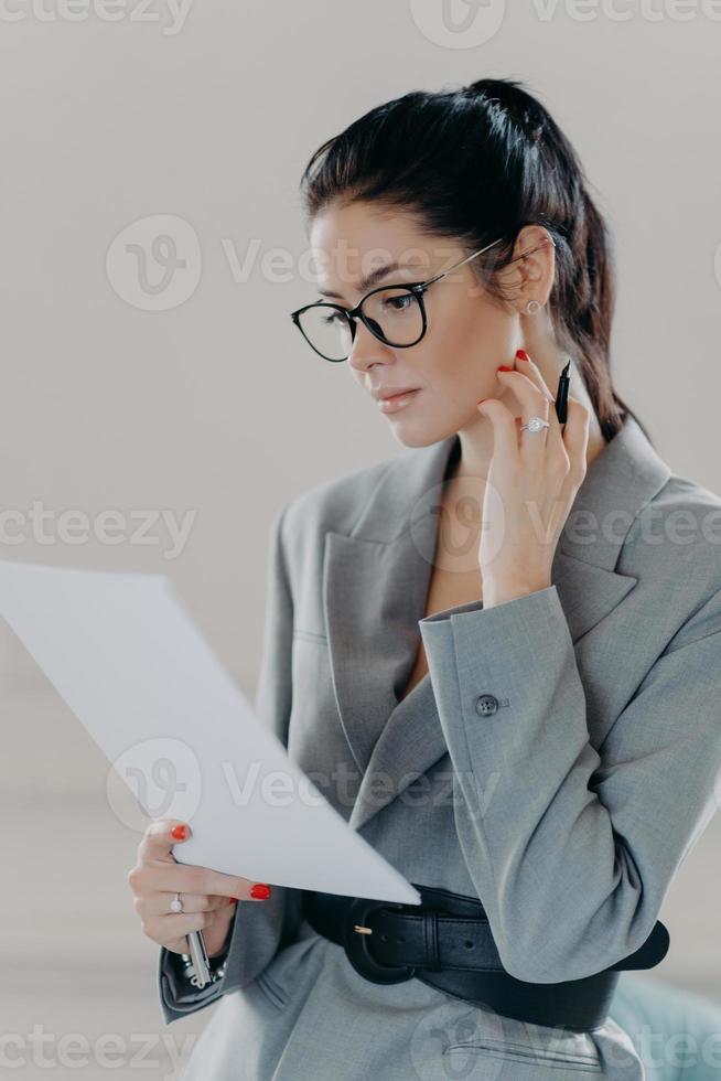 Successful female executive manager looks through paper documents, analyzes data, studies contract information, holds pen, wears spectacles, formal outfit, reviews monthly report or employee resume photo