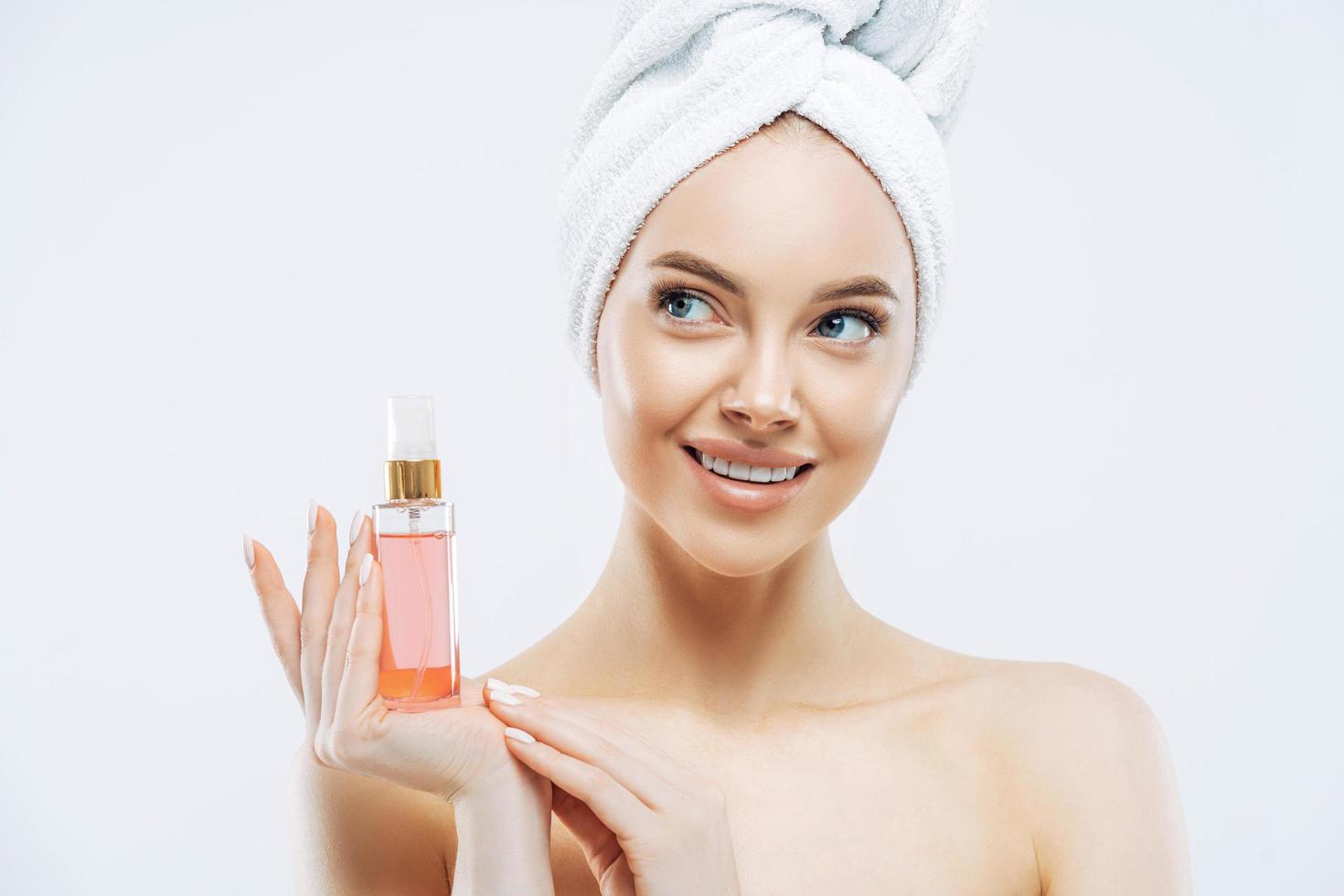 Beauty portrait of happy smiling European woman with healthy skin, makeup, holds bottle of aromatic parfume, stands naked indoor, wears towel on head. Women, cosmetology and skin care concept photo