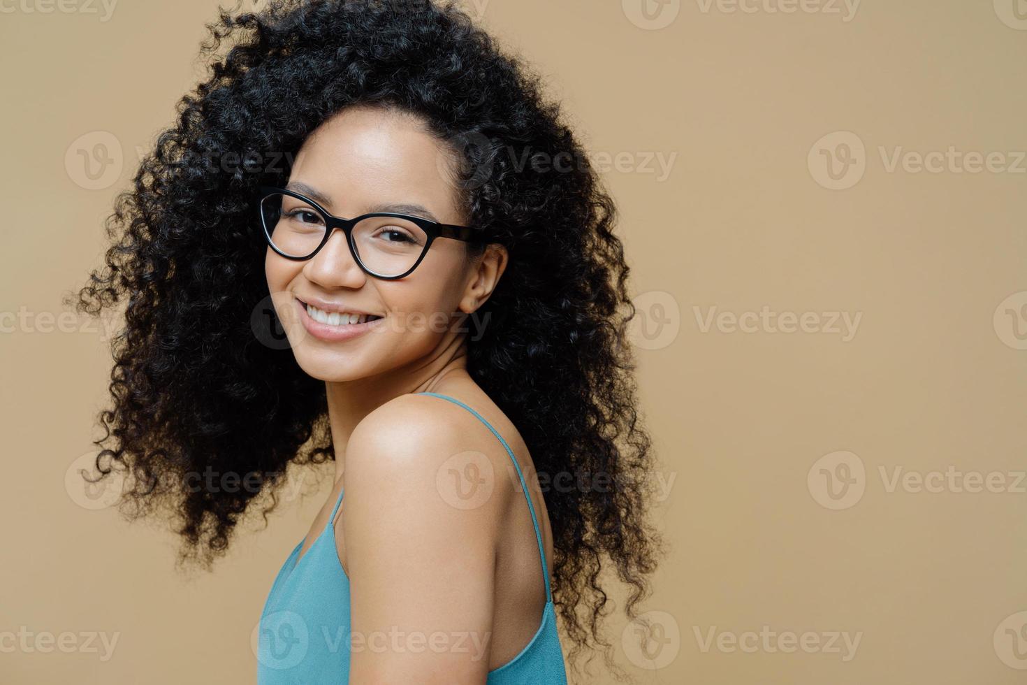 Magnetic pretty dark skinned woman stands sideways indoor, wears dress, shows bare shoulders, smiles gently, has curly bushy hair, looks through optical glasses at camera, isolated on beige wall photo