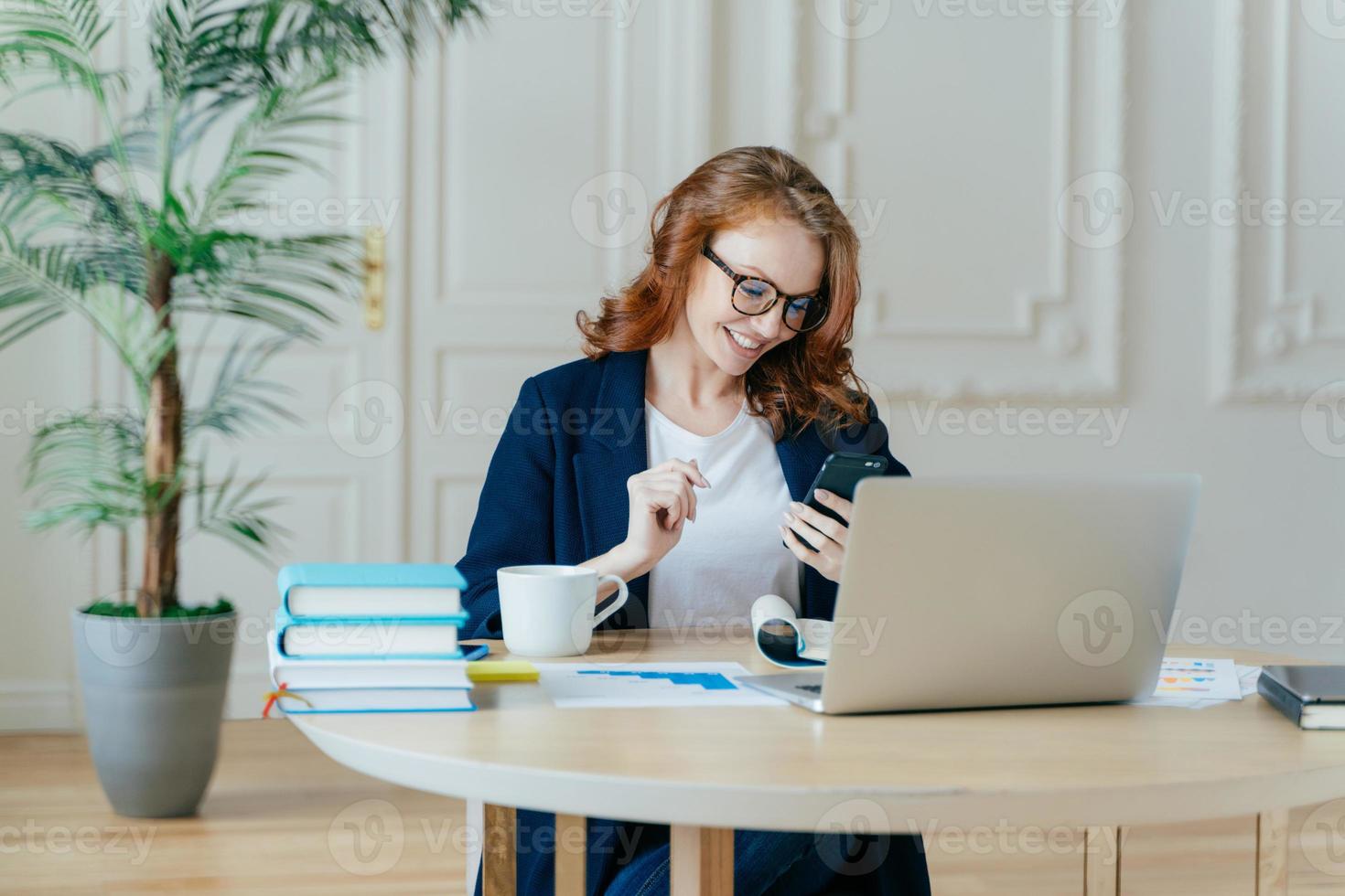 mujer pelirroja sonriente enfocada en un celular moderno, feliz de recibir mensajes de texto, posa en el lugar de trabajo, bebe café, trabaja en un proyecto financiero y se prepara para una videoconferencia con un socio comercial foto