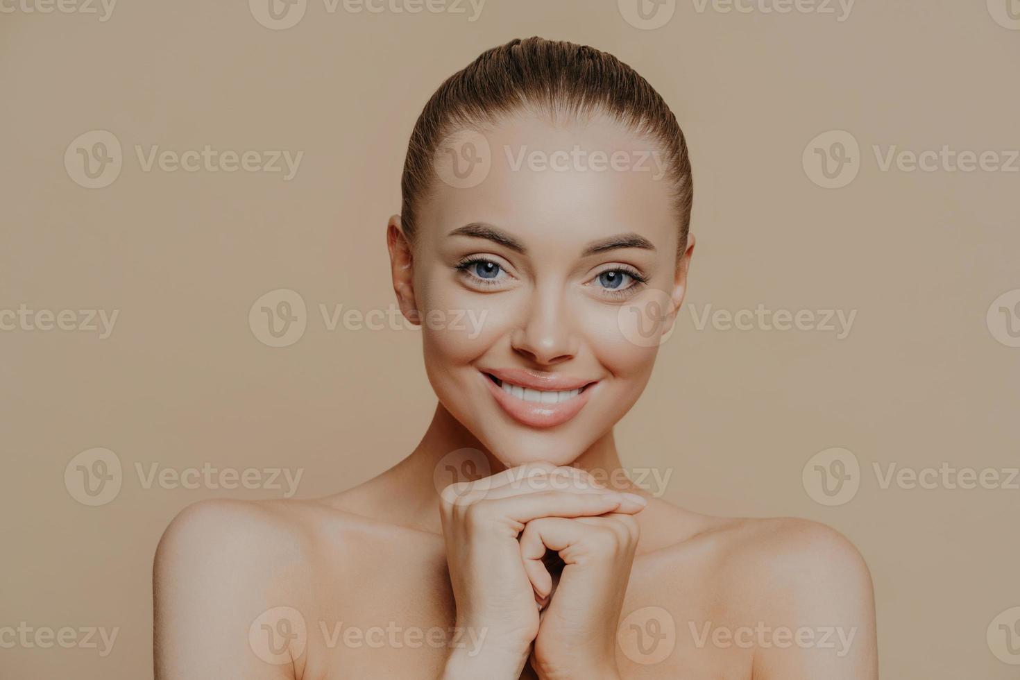 Close up shot of adult woman with fresh daily makeup, smiles toothily and keeps hands under chin, stands half naked indoor, feels refreshed after cosmetic procedures. Beauty and face care concept photo