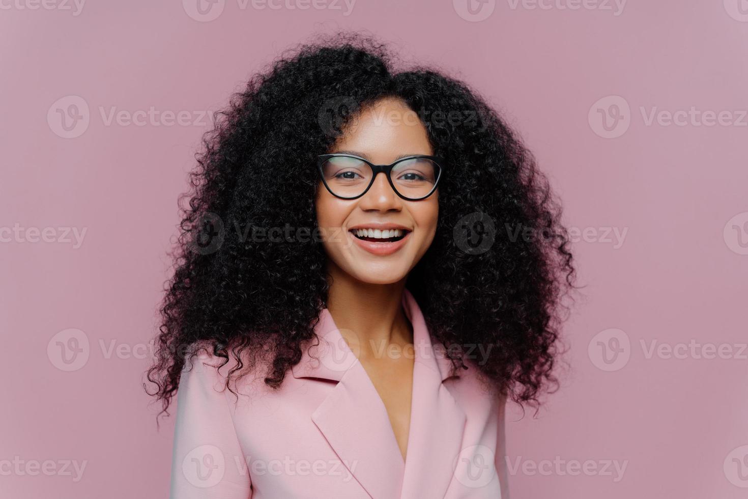 Close up portrait of happy ethnic young woman with crisp hair, looks through optical glasses, wears formal outfit, being in good mood, comes on work, happy get praise from boss, models indoor photo