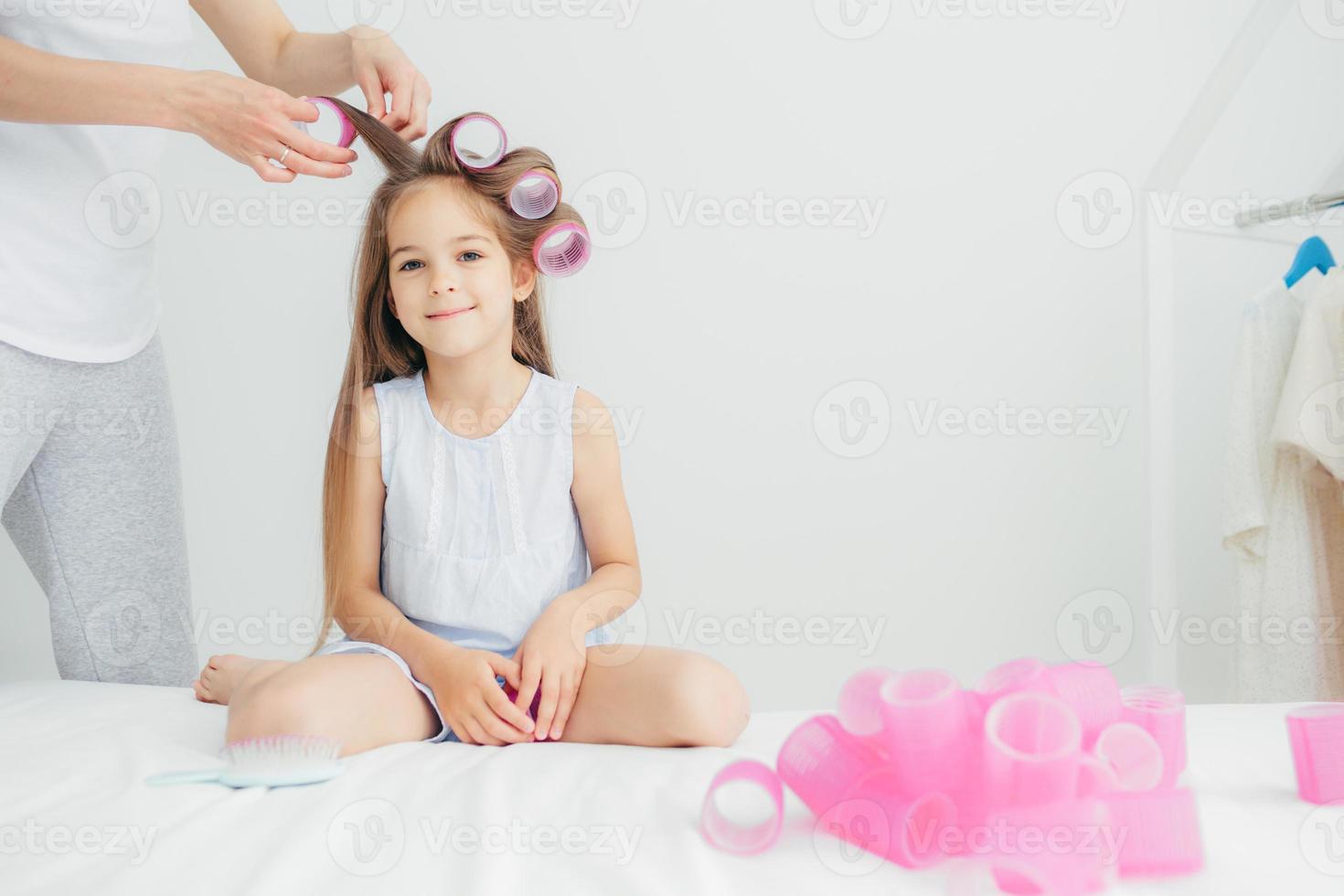 una foto horizontal de una linda hija pequeña se sienta cerca de su madre que enrolla rulos, posa contra un fondo blanco, se prepara para un evento festivo en el jardín de infantes. hermoso niño encantado tiene el pelo largo