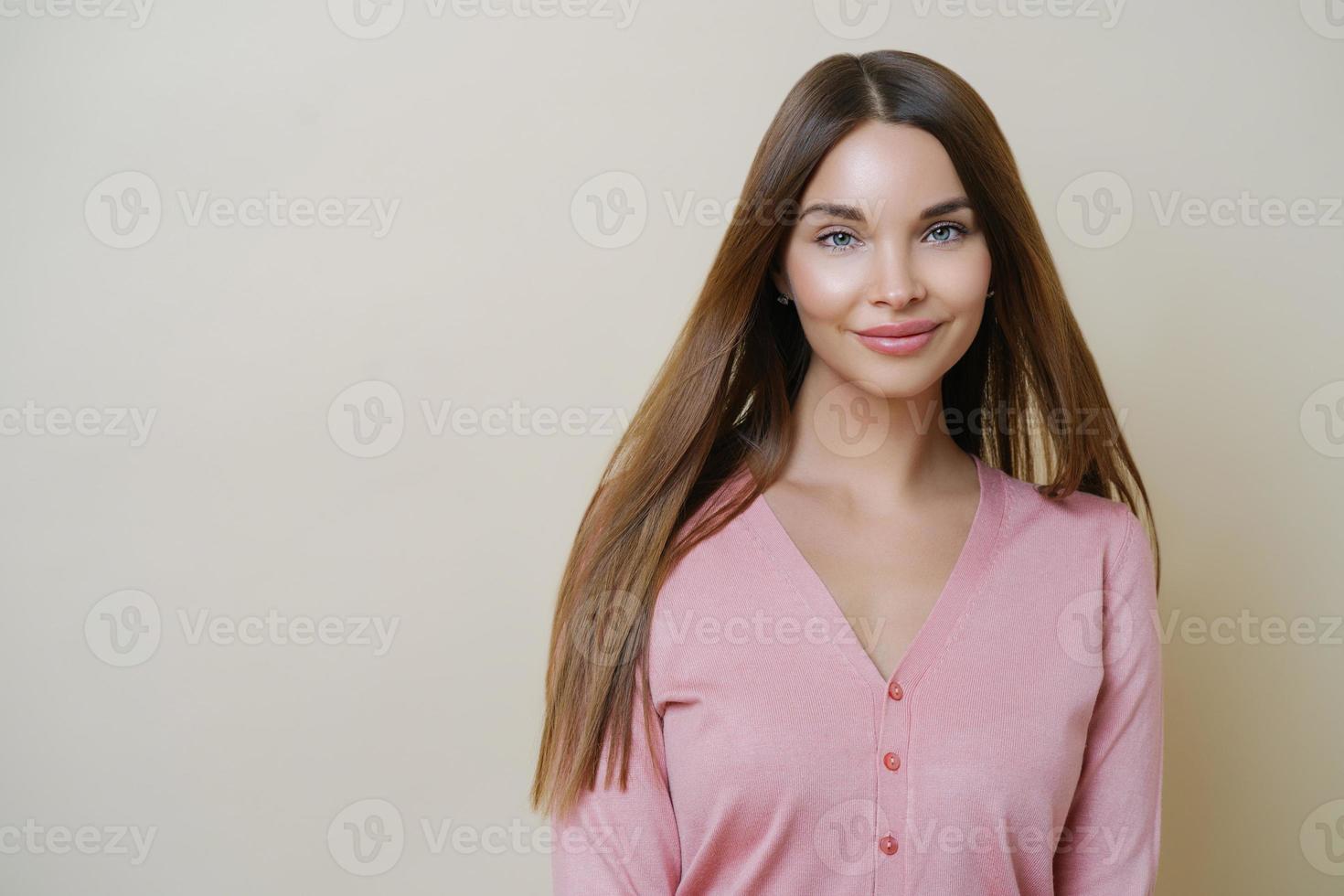 Horizontal portrait of charming European woman smiles gently at camera, has long straight hair, wears casual rosy sweater, going to have outdoor walk, stands against beige studio background. photo