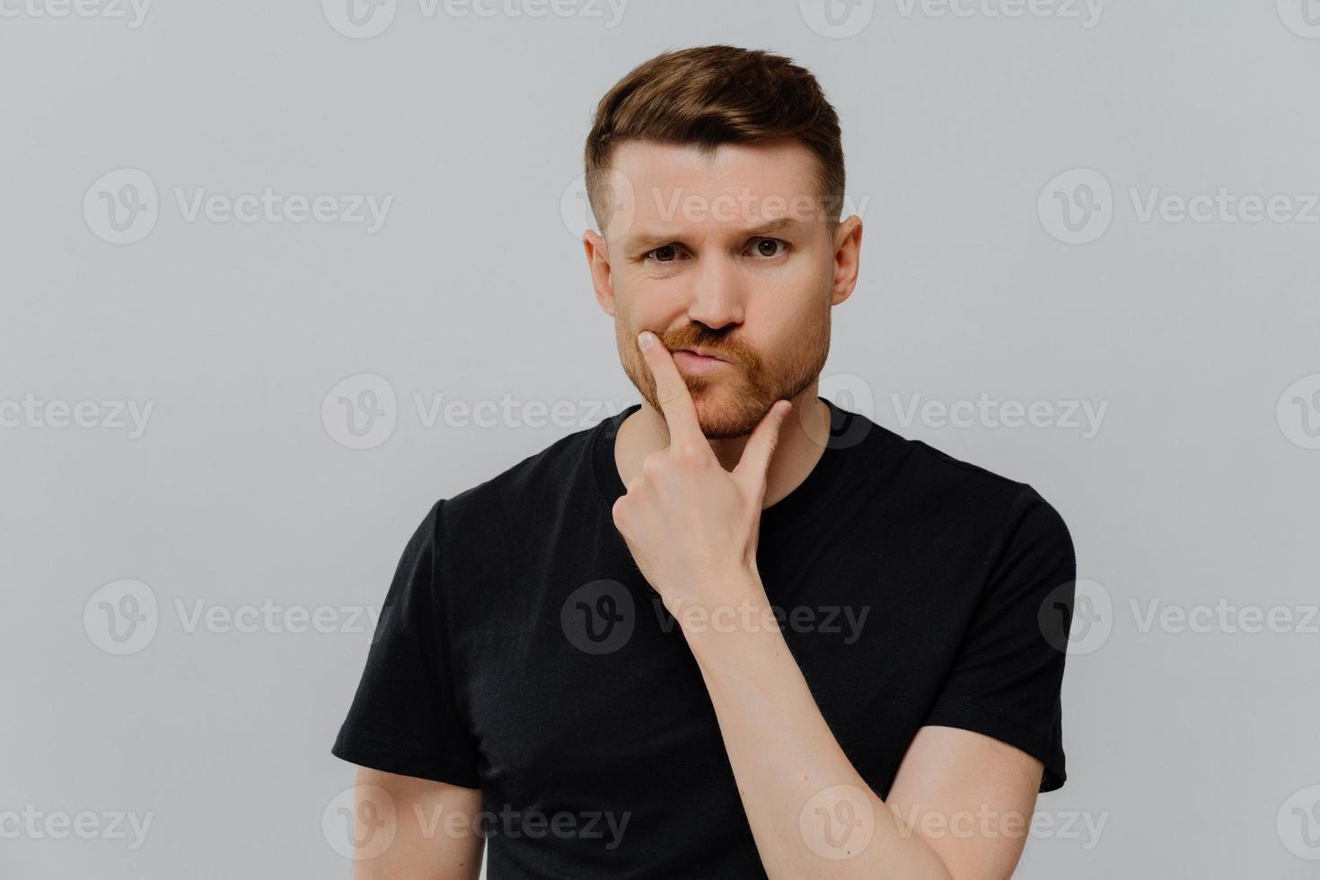 Studio shot of serious displeased man holds chin purses lips looks with thoughtful expression poners on something tries to make up his mind dressed in casual clothes isolated over blue background photo