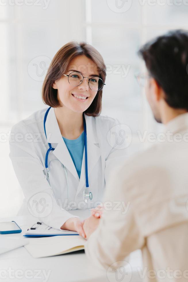 Medical trust concept. Vertical image of female doctor reassures her male patient, hold hand and talk calmly about illness cure, gives professional consultation and support, pose at doctors office photo