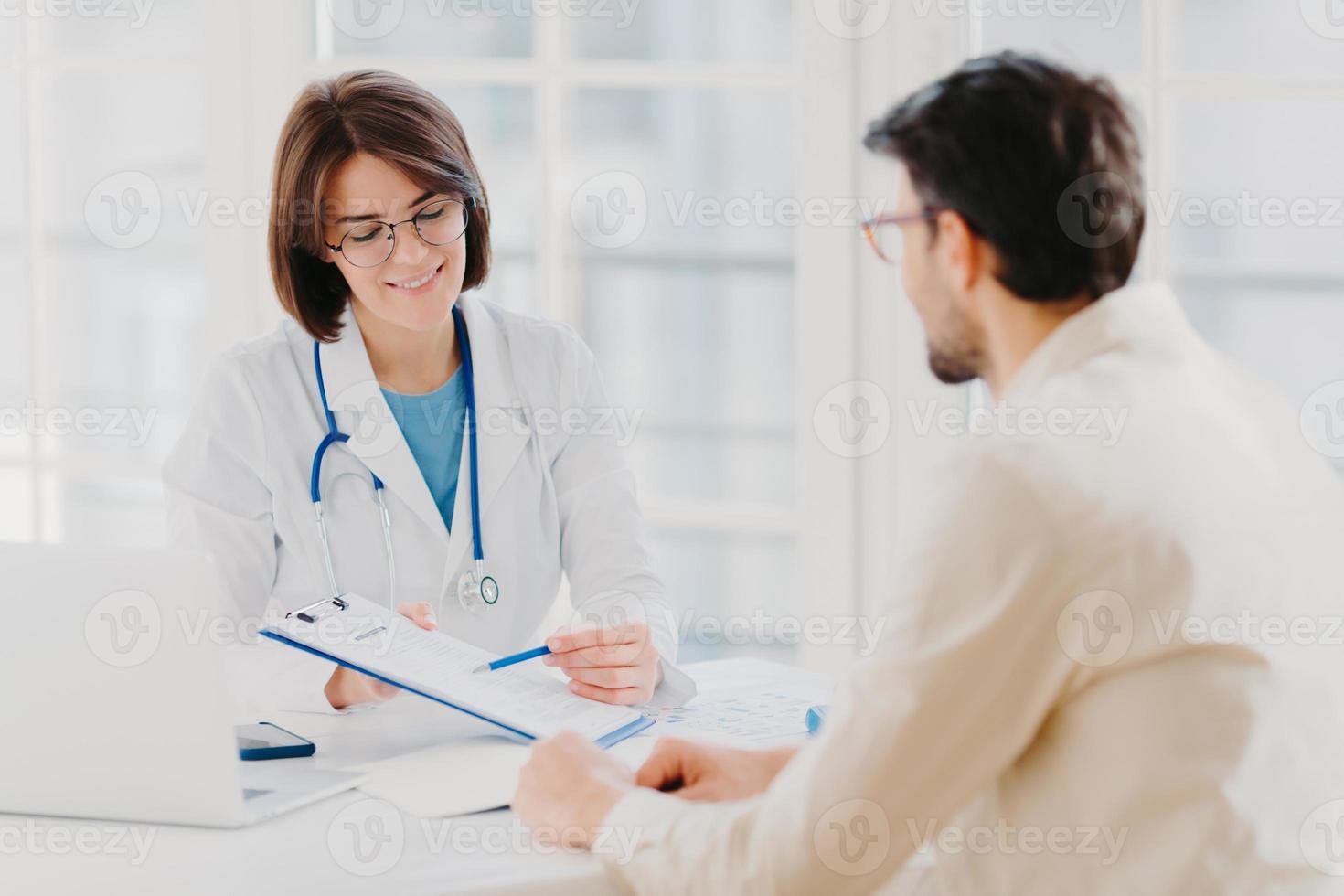 Medical appointment. Female doctor gives professional medical help to male patient, explains written information on paper in clipboard, gives support and good service, pose at hospital near desktop. photo