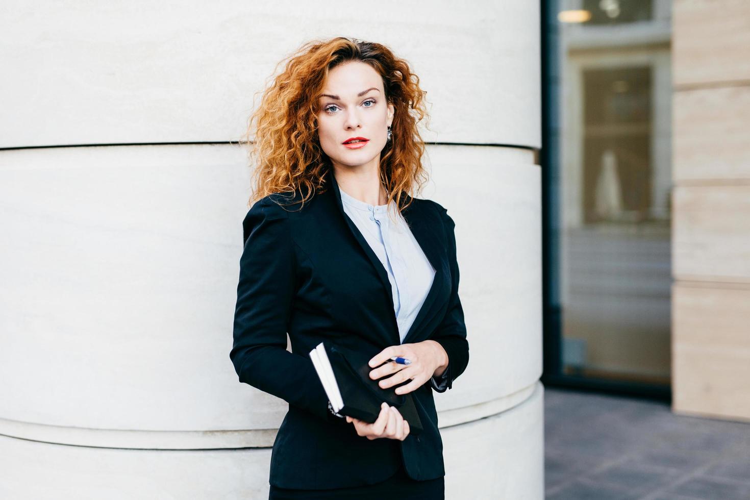 Young beautiful woman wearing formal clothes, dressed formally, holding pocketbook with pen, having bushy hairstyle, standing near office builduing. Successful pretty female office worker working photo