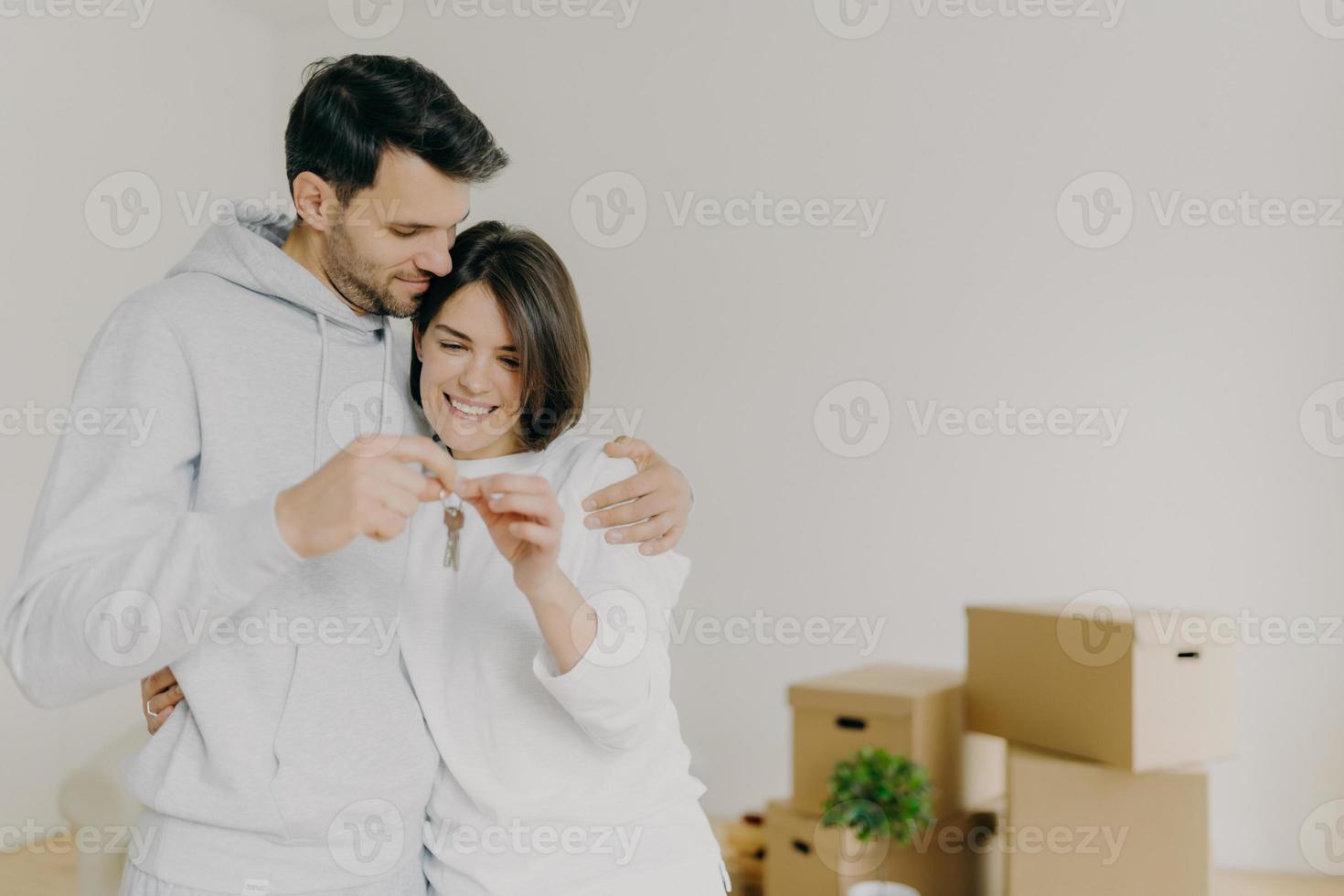 los propietarios de casas encantados de mujeres y hombres disfrutan de comprar un piso nuevo, tienen las llaves de su propio apartamento, posan contra una pared blanca con cajas de cartón desempaquetadas. hipoteca familiar, propiedad, bienes inmuebles. foto