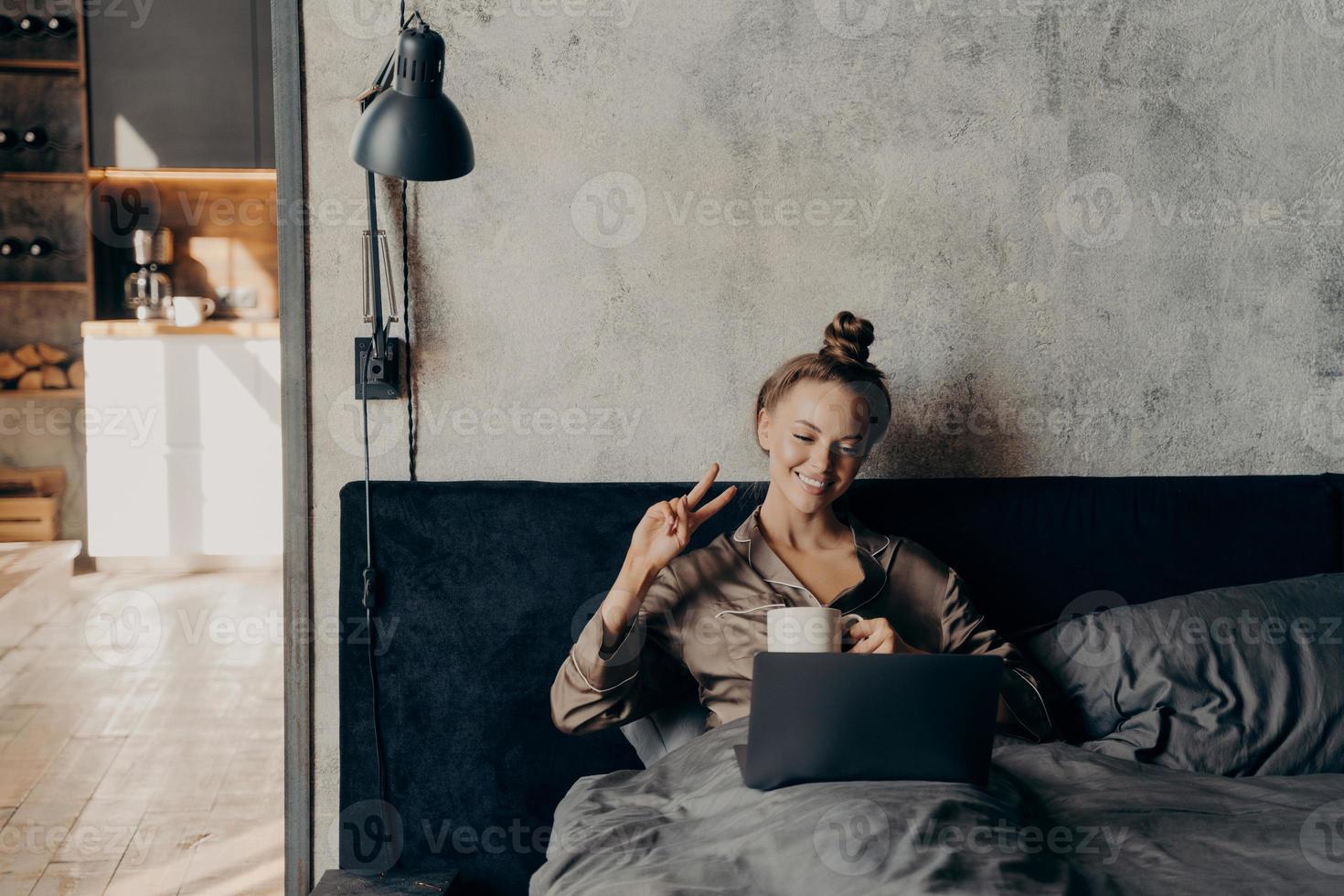 Lovely joyful young girl lying in bed with cup of coffee and having video call with friends online on laptop photo