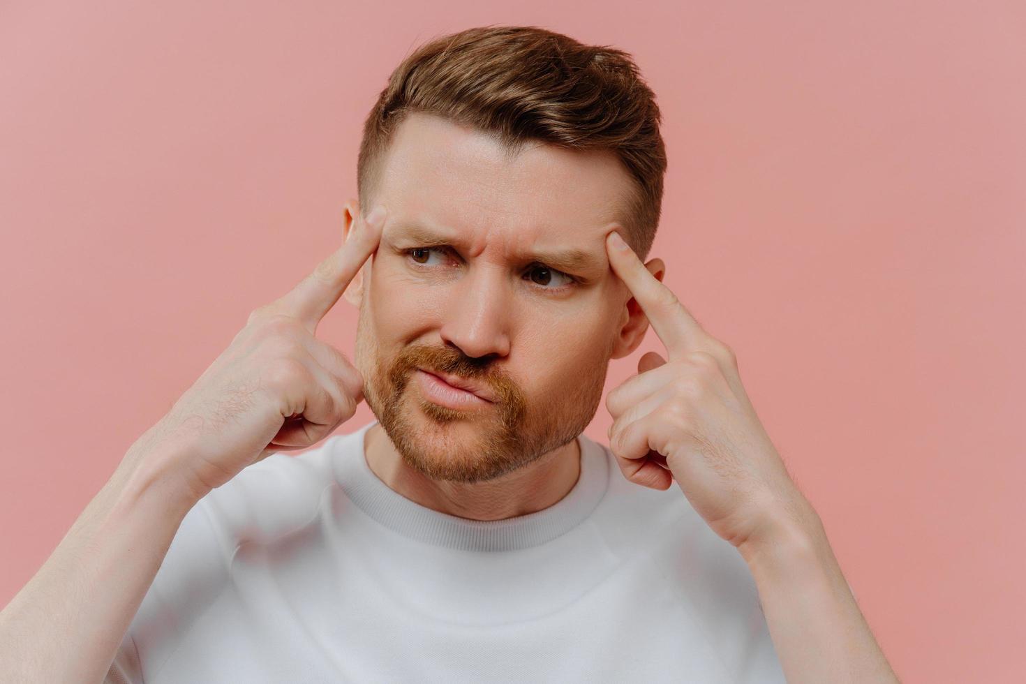 Close up shot of serious bearded adult man keeps fingers on temples concentrated aside tries to recall something in mind dressed casually isolated over pink background. Male model thinks deeply photo