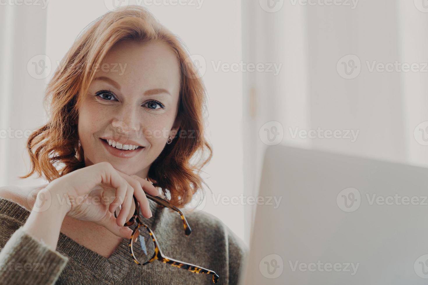 Pretty ginger businesswoman searches online web informationn via laptop computer, has pleasant smile, holds optical glasses, sits at coworking desktop, busy with freelance work. Technology concept photo