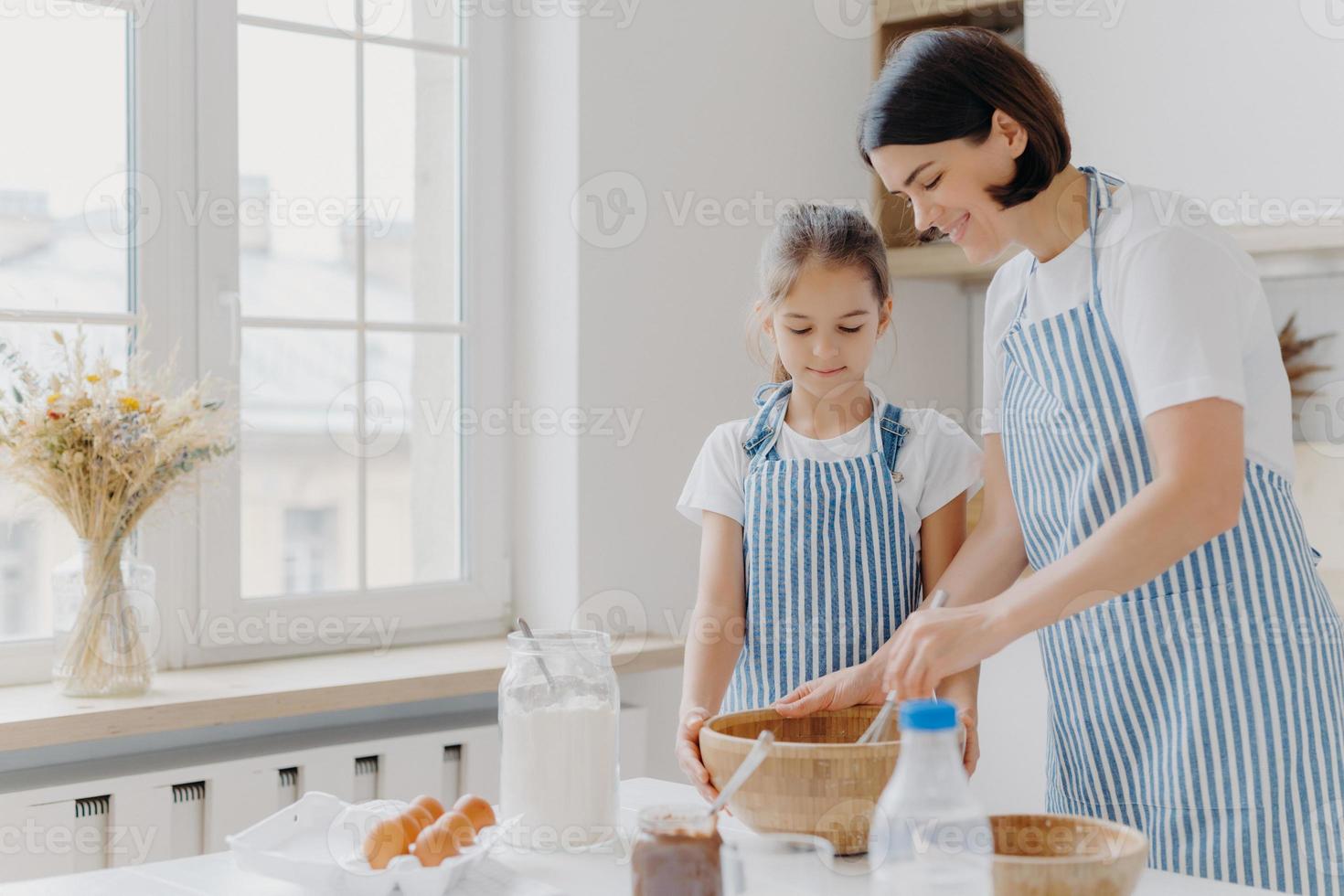 foto de mujer morena con delantal a rayas, mezcla ingredientes con batidor, le muestra a su hija pequeña cómo cocinar, parada en la cocina cerca de la mesa con productos frescos. madre e hijo ocupados preparando comida