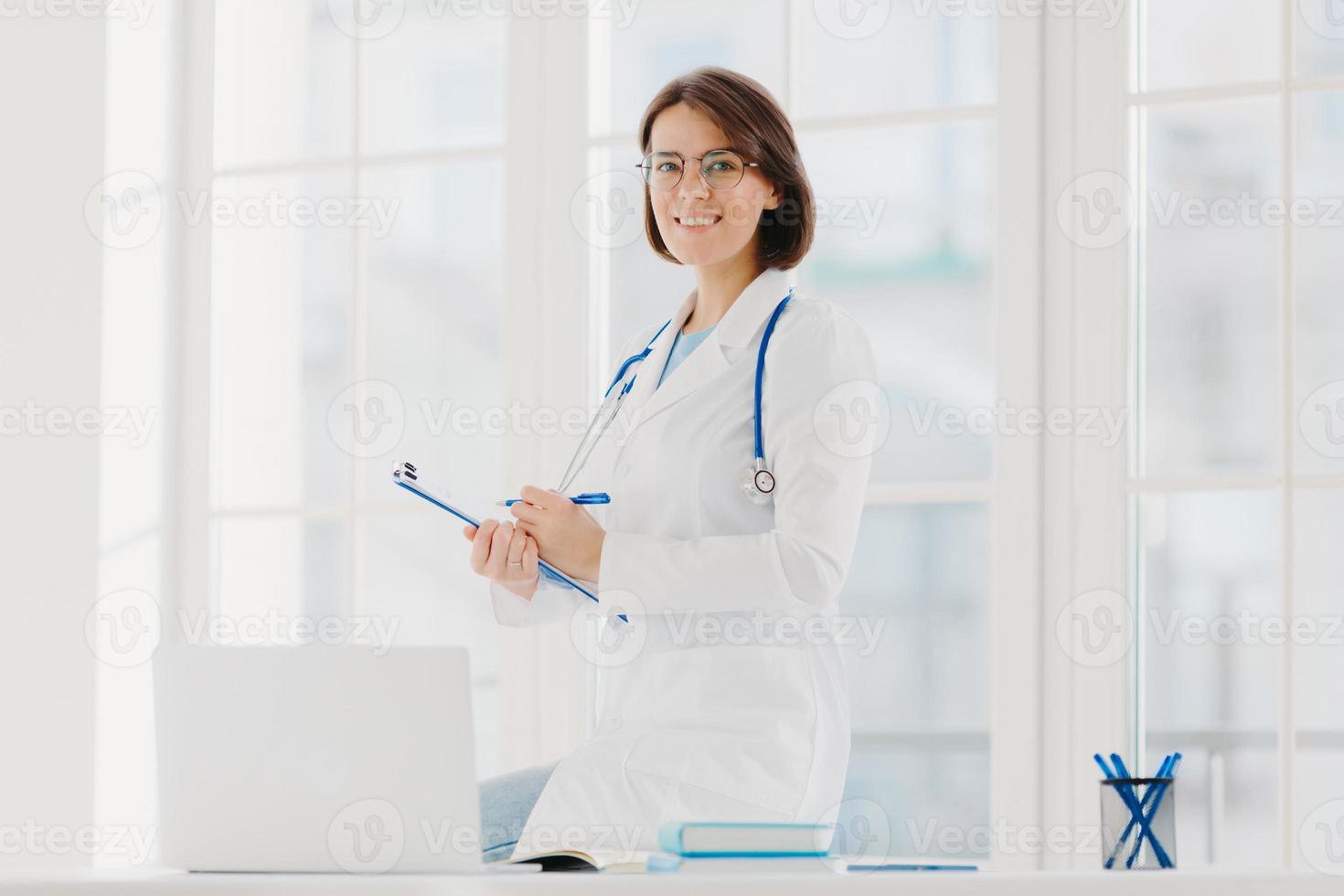 Indoor shot of professional female doctor supplies medic care assistance, poses at desktop with laptop, prepares document agreement paper offer, wears optical glasses and white medical gown. photo