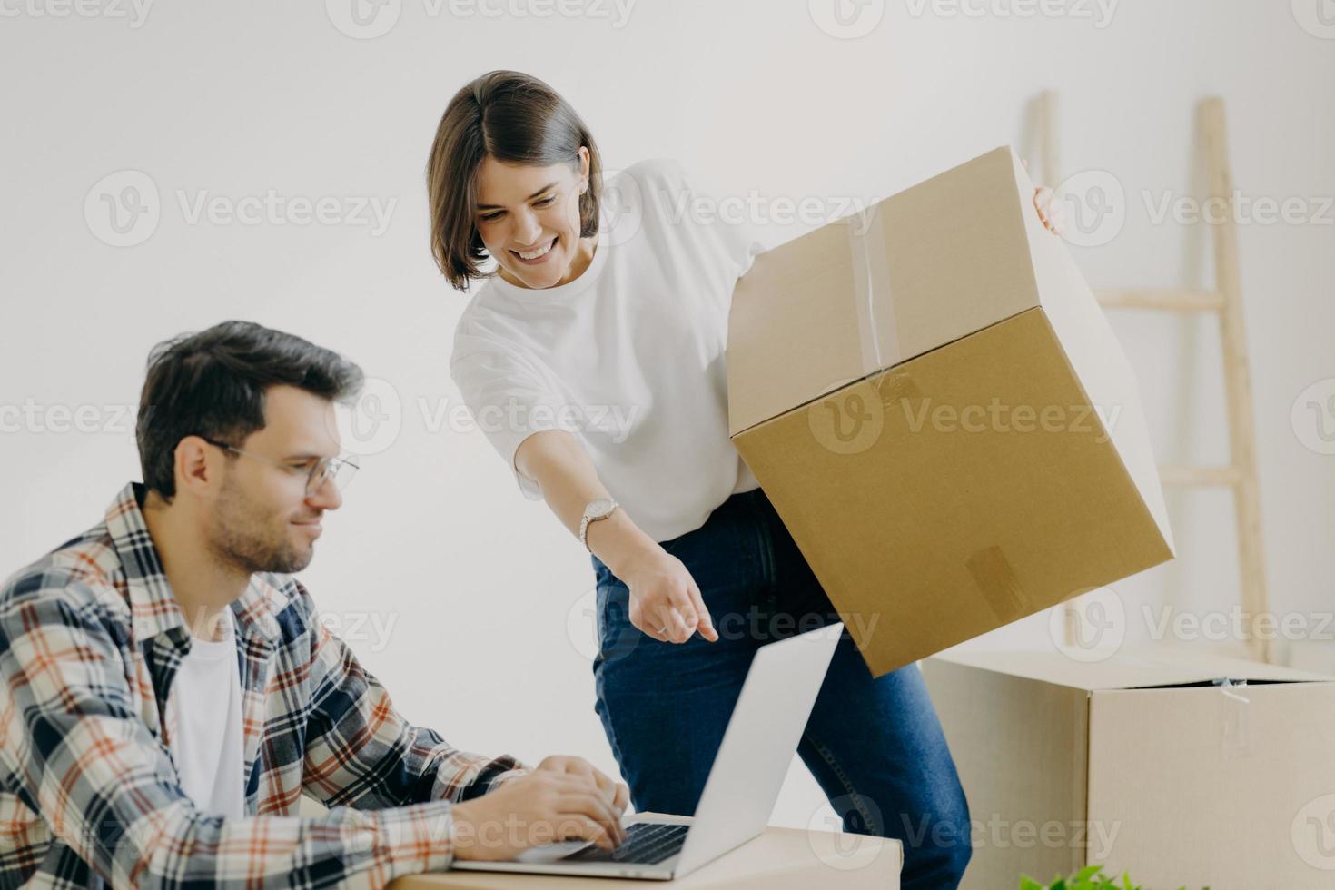 Indoor shot of happy family couple look attentively at laptop computer, search good moving company, carry personal belongings in big cardboard boxes, woman points into display of modern gadget photo