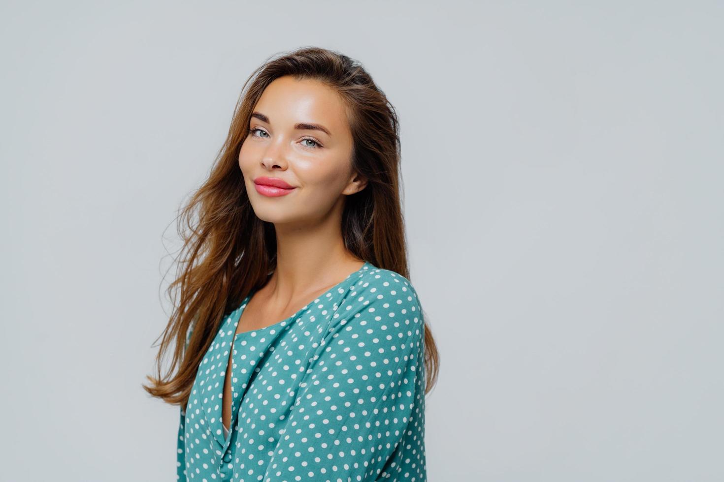 Profile shot of pretty pleased woman wears makeup, rosy lipstick, dressed in polka dot blouse, has healthy skin, poses against white background with blank space for your advert or promotion. photo