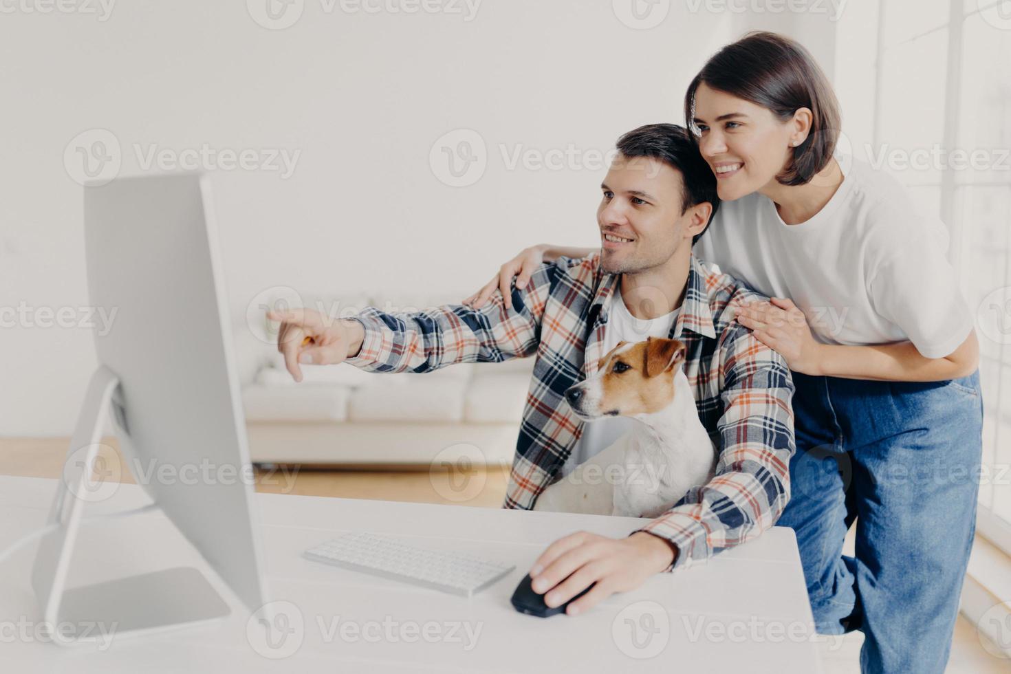 Happy man and his girlfriend consult about future project, point into monitor, pose at workplace together with dog in home interior, watch online tutorial video, spend time in modern apartment photo