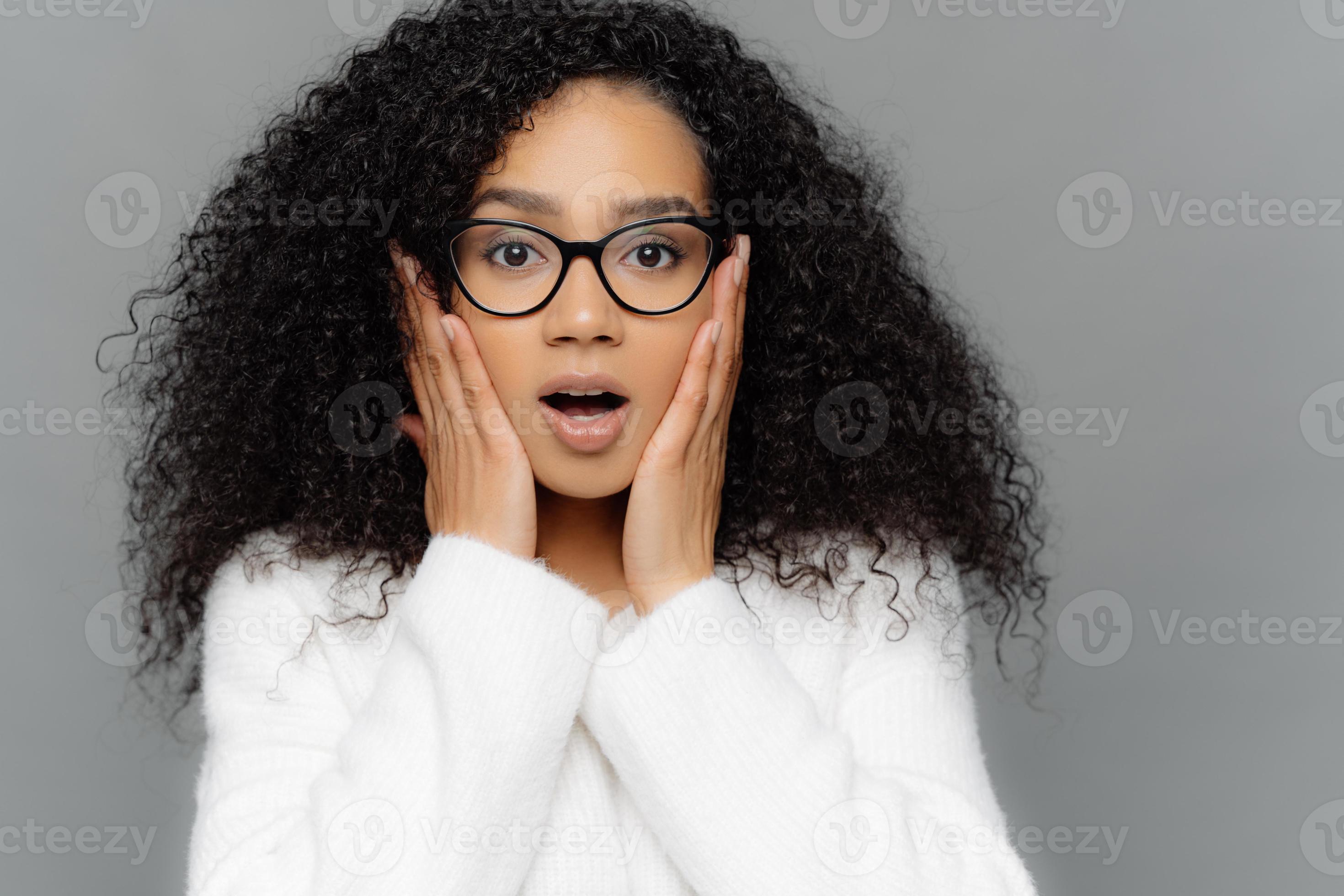 Close up portrait of shocked concerned young woman with dark skin and curly  hair, hears terrifying news, keeps hands on cheeks, wears spectacles and  white sweater, isolated on grey background 8358645 Stock