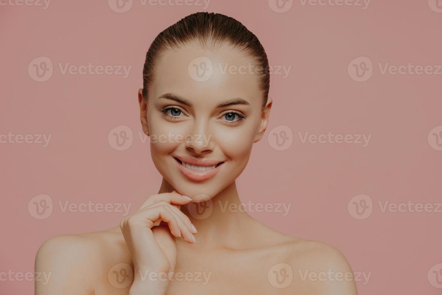 Studio shot of pretty charming woman touches chin, has healthy glowing skin, cares about complexion, stands naked indoor, has cosmetology procedure, isolated on pink background. Beauty concept photo