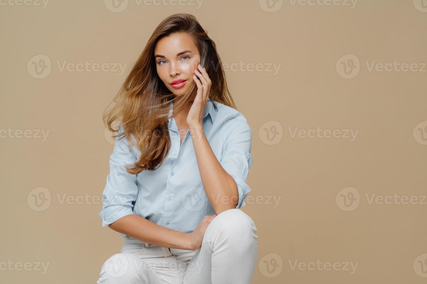 Pretty female feels relaxed while sits at chair, dressed in stylish shirt and white trousers, poses over brown hair, copy space for your promotional content. Beautiful woman in fashionable clothes photo