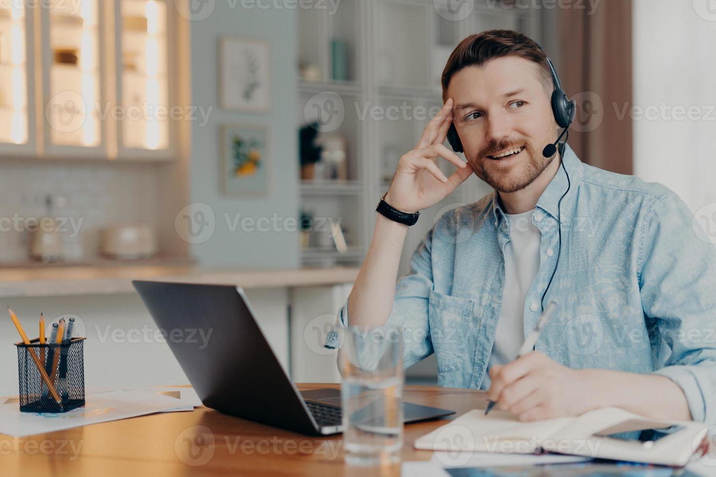 Pensive thoughtful man makes notes uses laptop computer headset writes down notes in notepad supplies customer support solves clients issues sits at desk in coworking space. Hotline manager. photo