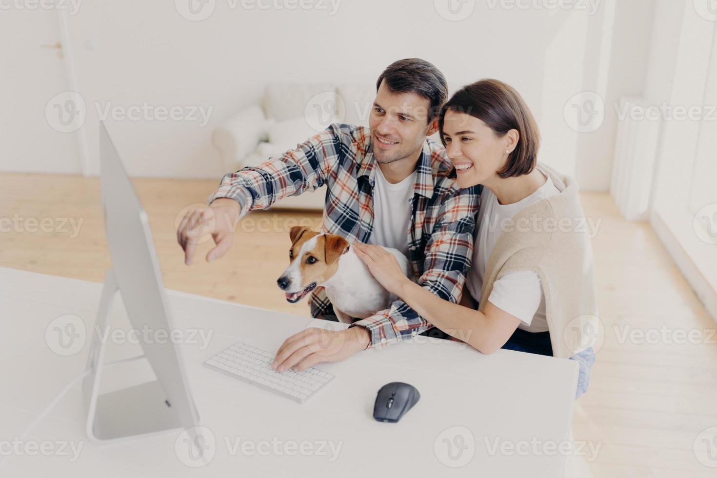 Overjoyed woman and man has happy looks, watch online movie at home together, play with dot, sit at white table, search information via wifi, dressed in casual clothes, Family couple browse internet photo