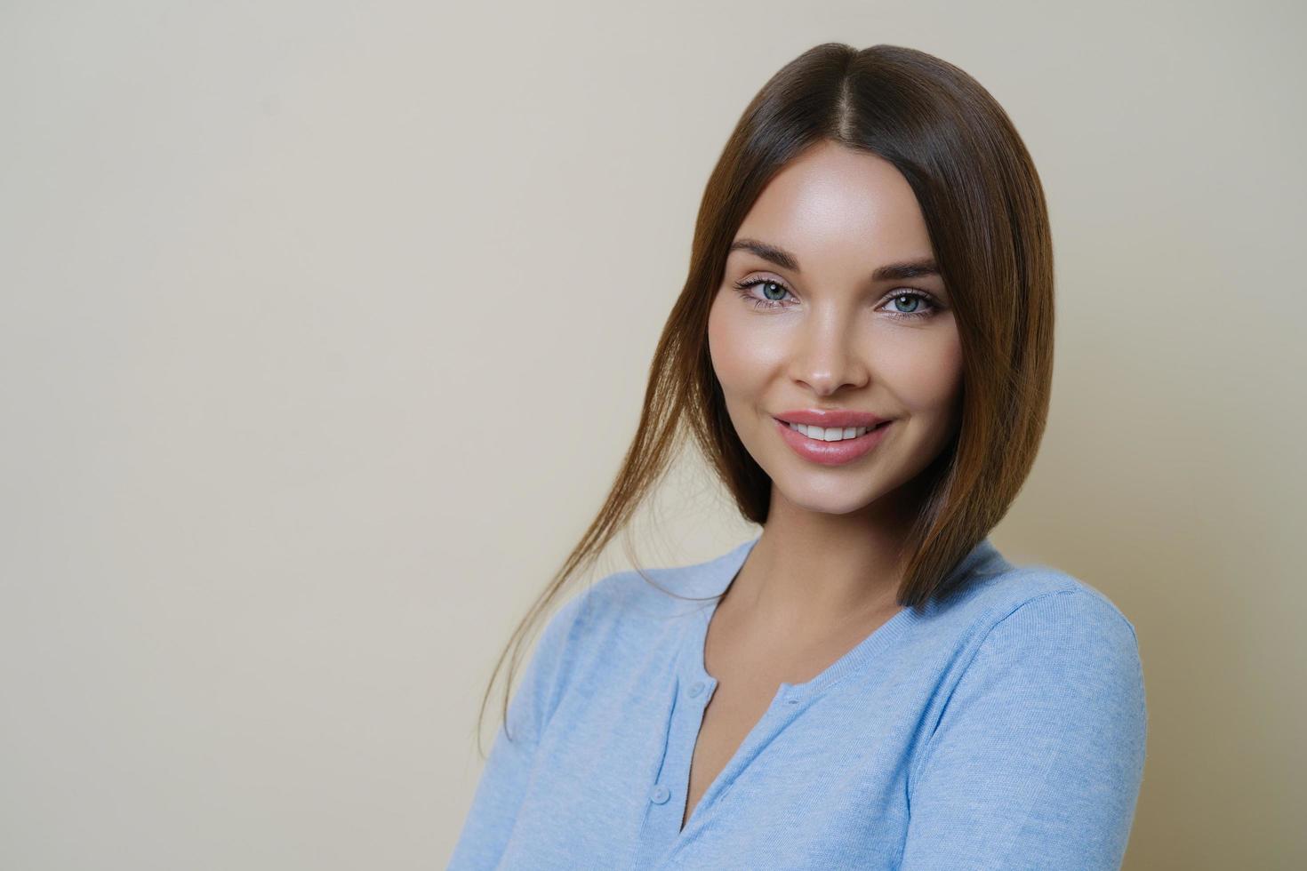 Close up portrait of beautiful lady with beauty face and healthy clean skin, smiles pleasantly, has shiny straight brown hair, looks confidenlty at camera, isolated over beige wall. Hair product, care photo