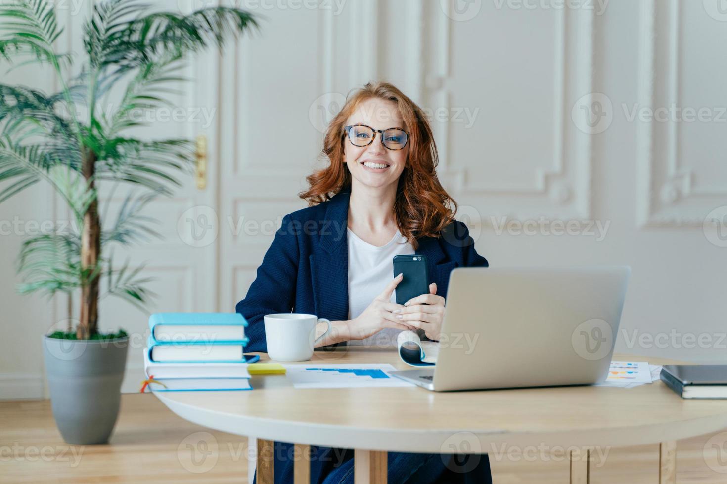 Horizontal shot of optimistic woman installs application on modern cell phone, checks notification, surrounded with paper documents, works on laptop computer, has red hair, dressed formally. photo