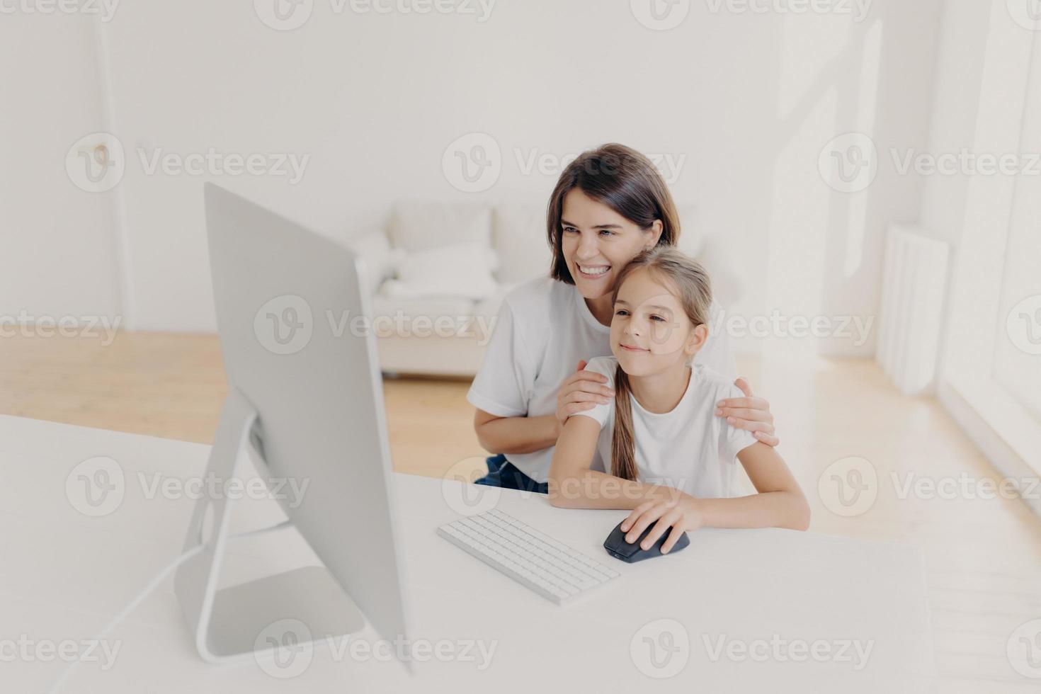 Indoor shot of happy lovely mother and daughter watch funny cartoon on computer, have pleasant smiles, enjoy togetherness, spend free time or weekend at home, spacious light empty living room photo