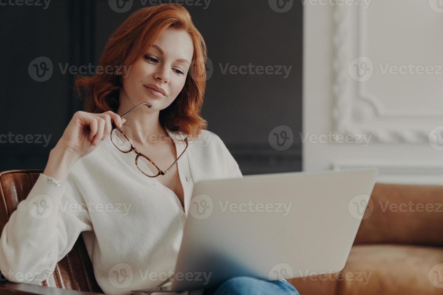 Concentrated serious redhead woman makes remote job indoors, sits in comfortable armchair, holds transparent glasses, reads publication on internet website, uses home wifi. Working time, technology photo