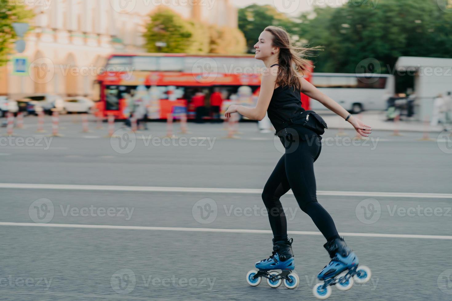 una foto al aire libre de una joven activa y delgada disfruta del patinaje sobre ruedas durante el tiempo libre vestida con poses de ropa negra activa en un lugar urbano en la carretera contra un fondo borroso con transporte. concepto de afición