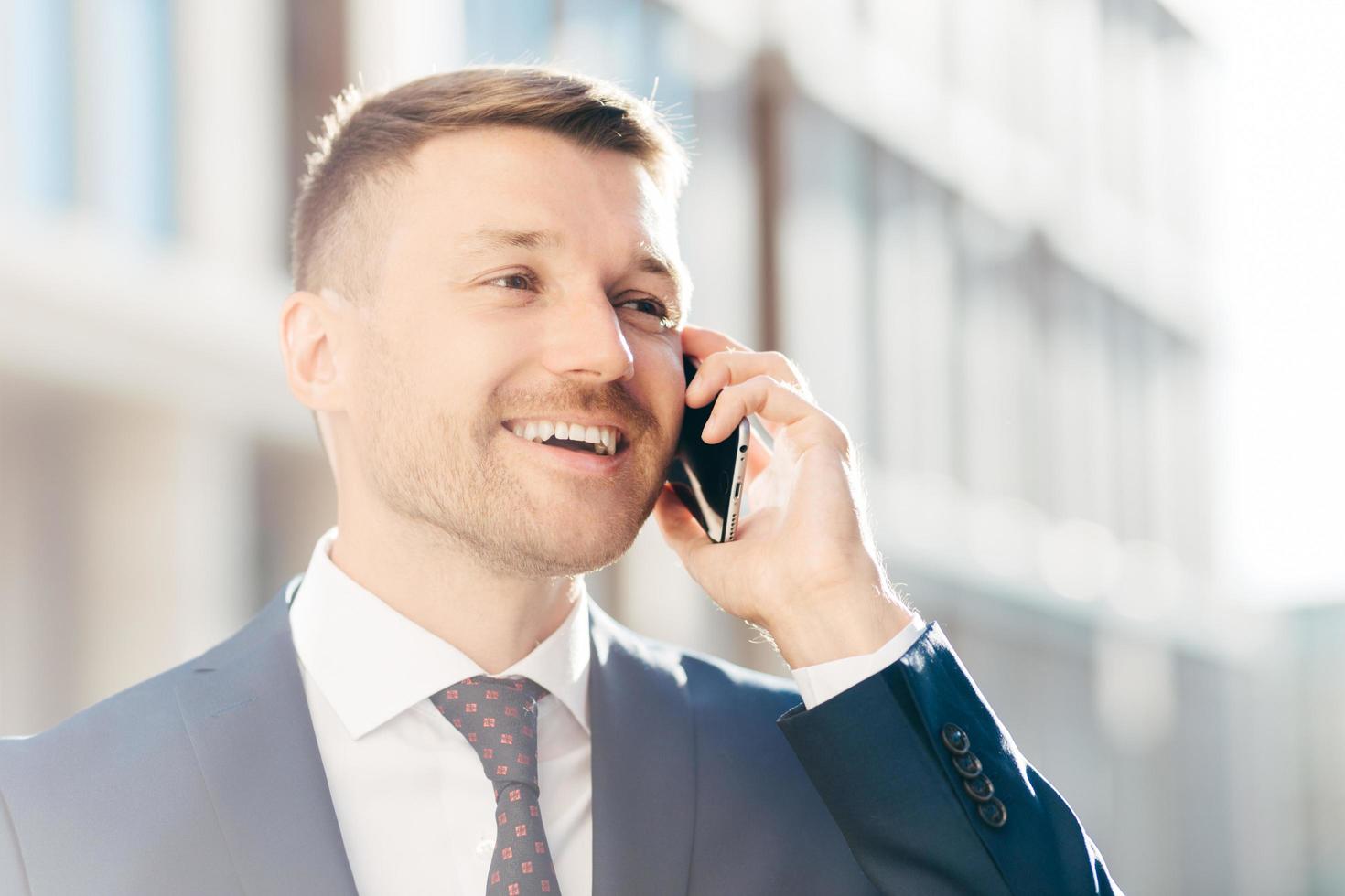 Portrait of successful positive office employee has phone conversation, looks happily into distance, discusses money payment. Male executive manager in formal suit speaks with client via cellular photo